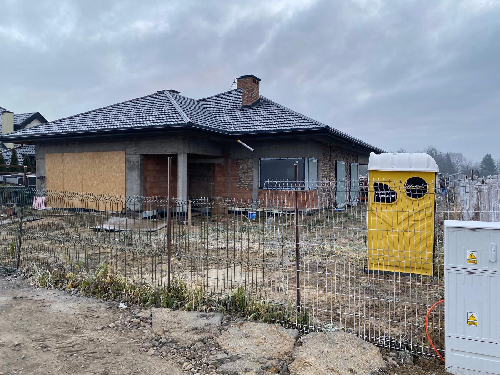 Dom jednorodzinny w trakcie budowy, z widoczną niezabudowaną przestrzenią garażową zabezpieczoną płytą OSB, ogrodzony siatką, z przenośną toaletą na terenie budowy w pochmurny dzień.