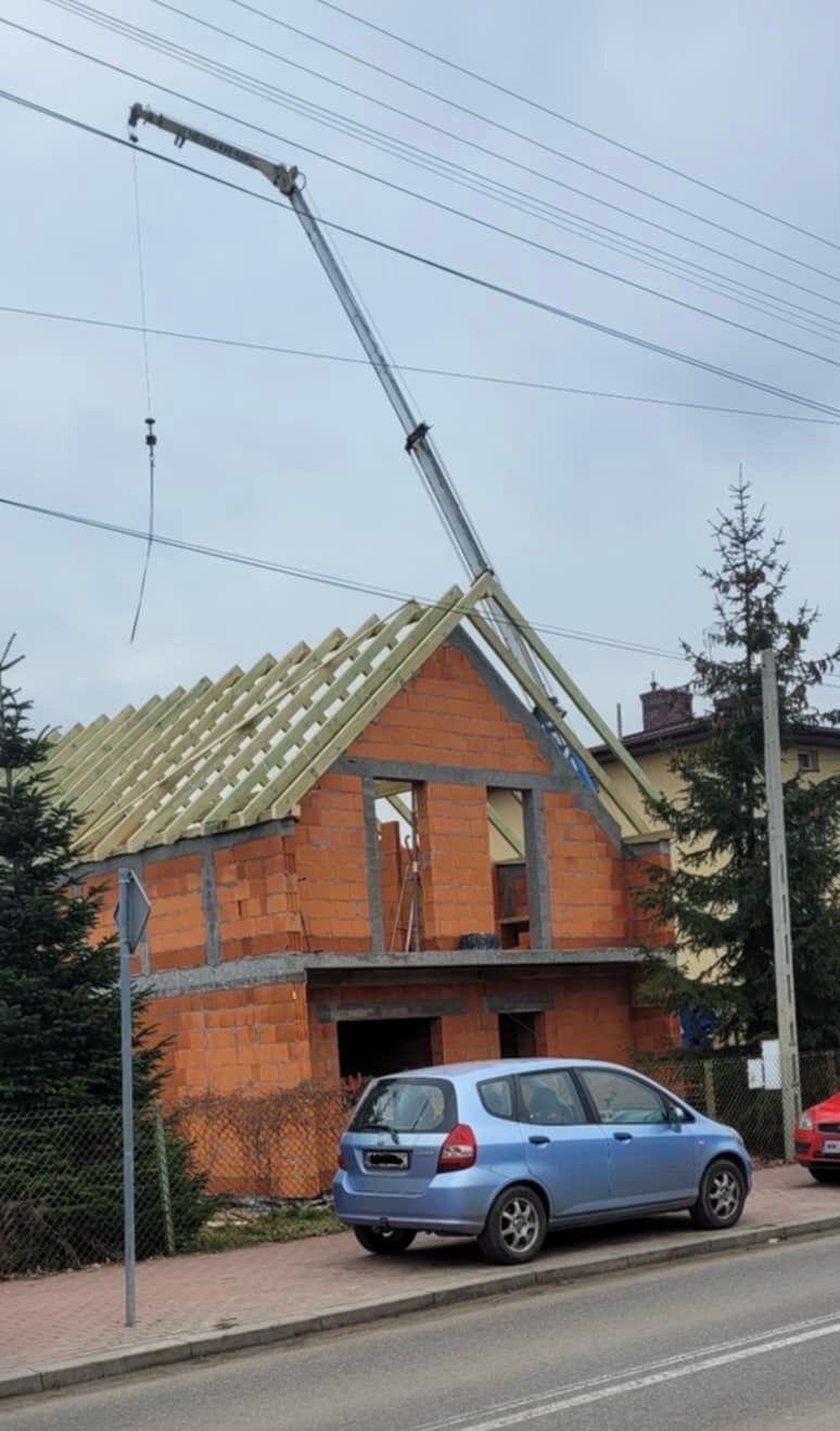 Budowa domu jednorodzinnego w stanie surowym otwartym, z widocznym dźwigiem na tle pochmurnego nieba, niebieski samochód osobowy zaparkowany przed budynkiem.