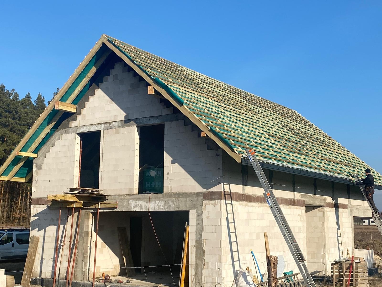 Budowa domu jednorodzinnego w trakcie realizacji: murowane ściany z betonu komórkowego, drewniana konstrukcja dachu pokryta zieloną membraną, drabina oparta o budynek, pracownik na dachu.