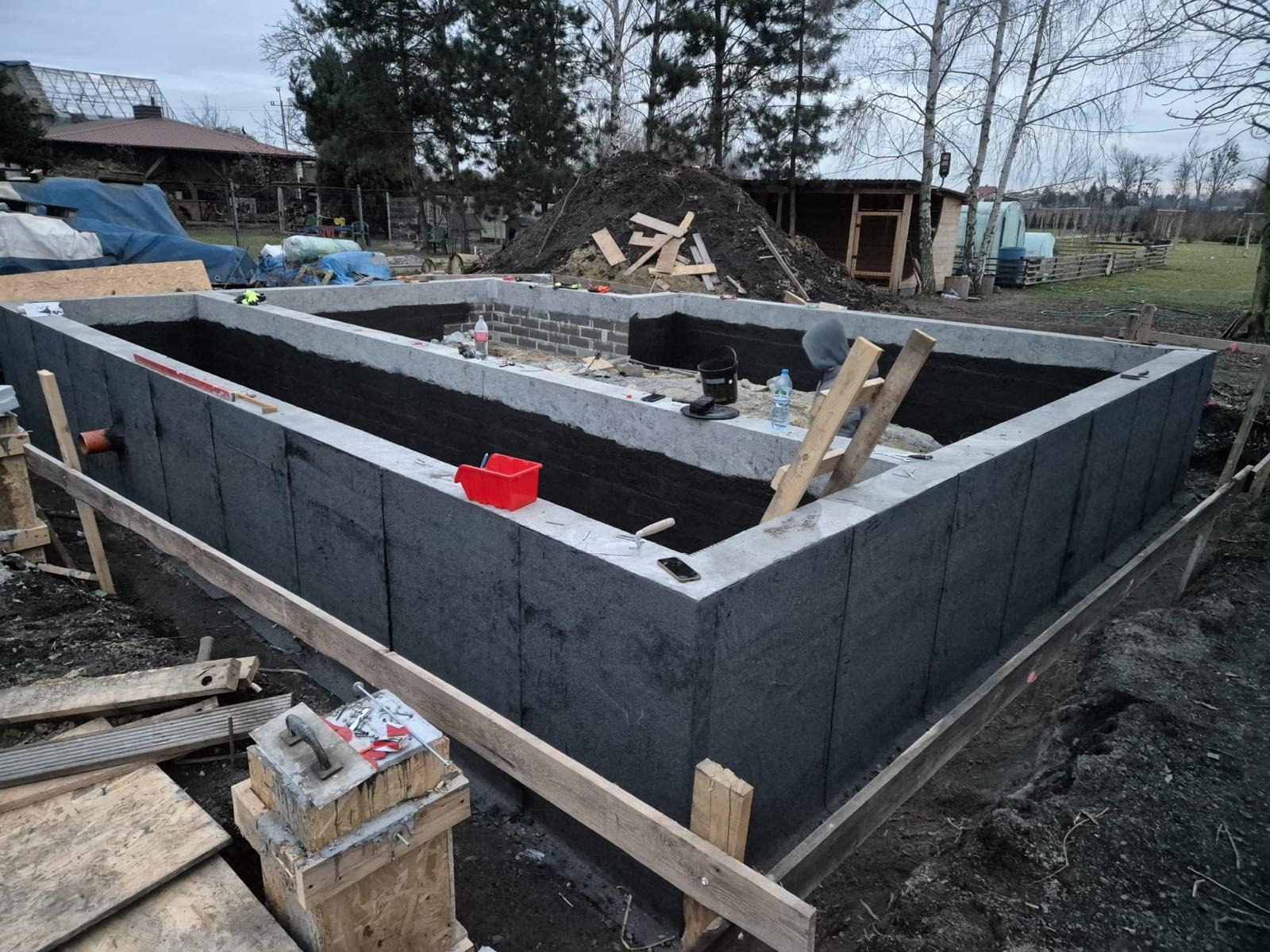 Betonowe fundamenty domu z izolacją pionową, widoczny fragment muru z bloczków betonowych, drewniane szalunki i narzędzia budowlane w otoczeniu działki.