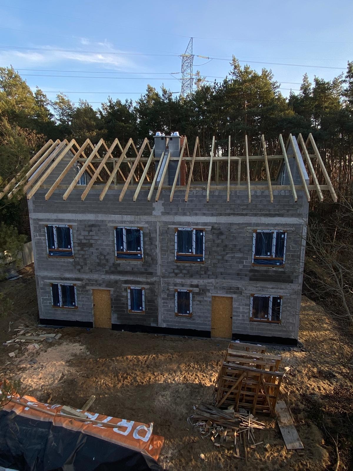 Surowy stan dwukondygnacyjnego domu z bloczków betonowych z drewnianą konstrukcją dachu w trakcie budowy, widoczne okna zabezpieczone folią, w tle las i słup wysokiego napięcia.