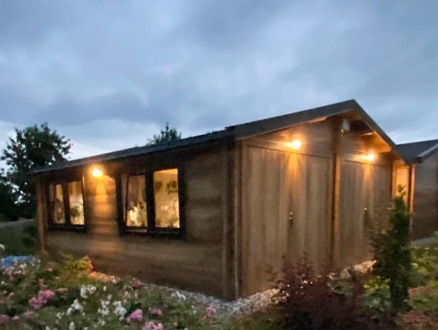 Drewniany domek ogrodowy z oświetleniem zewnętrznym, widoczny wieczorem na tle zachmurzonego nieba.