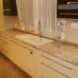 Beżowy blat kuchenny z delikatnym użyleniem, zintegrowany z białym zlewozmywakiem, nowoczesne szafki z podłużnymi uchwytami, drewniana podłoga i okno z jasną zasłoną w tle.