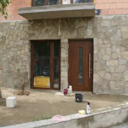 Wykończenie elewacji budynku kamieniem naturalnym w trakcie prac, widoczne narzędzia i materiały budowlane, okno z tablicą informacyjną, brązowe drzwi wejściowe.