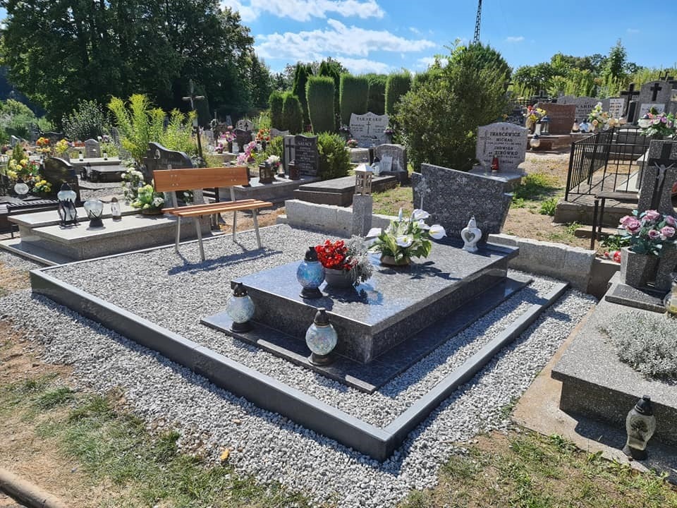 Nowy, ciemnoszary pomnik z polerowanego kamienia, otoczony jasnymi kamykami i obramowaniem, na cmentarzu z innymi nagrobkami i zielenią w tle, w słoneczny dzień.