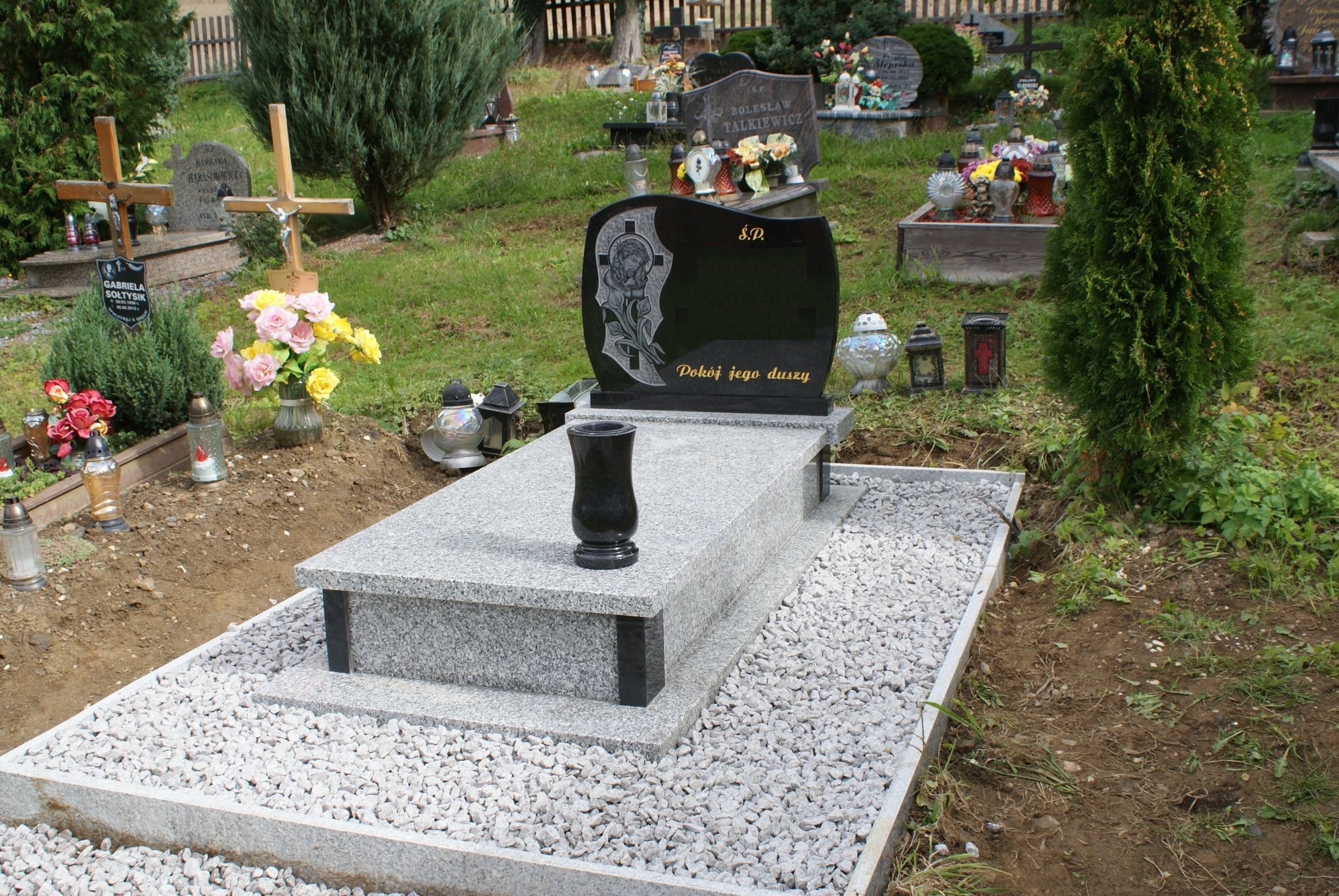 Nowo postawiony, jasnoszary pomnik z czarnym nagrobkiem, otoczony białym żwirem na cmentarzu, z widocznymi innymi grobami w tle.