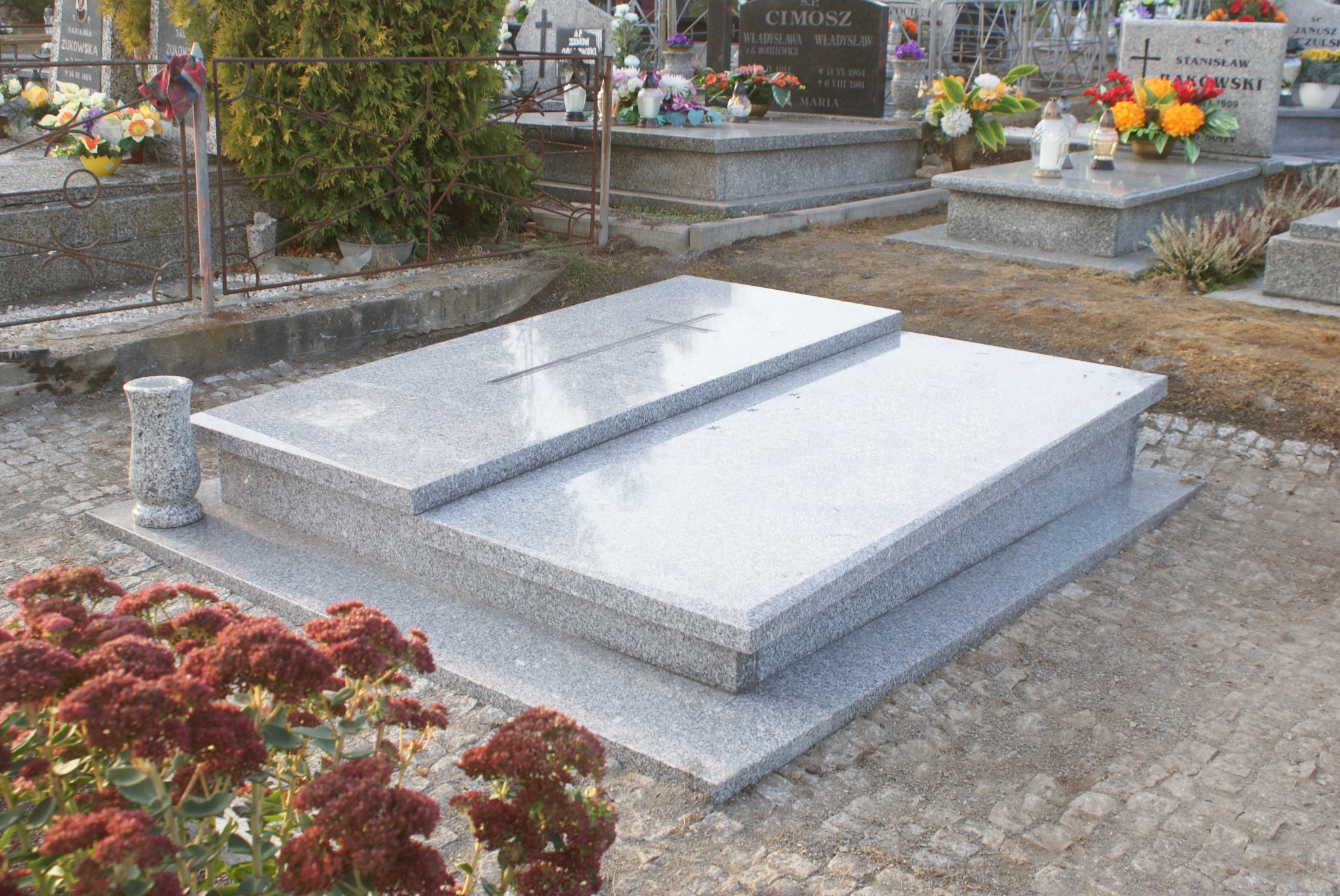 Jasnoszary, dwupoziomowy pomnik nagrobny z granitu z wyrytym krzyżem, widok z boku, z wazonem i czerwonymi kwiatami na pierwszym planie, w tle inne nagrobki i drzewa.