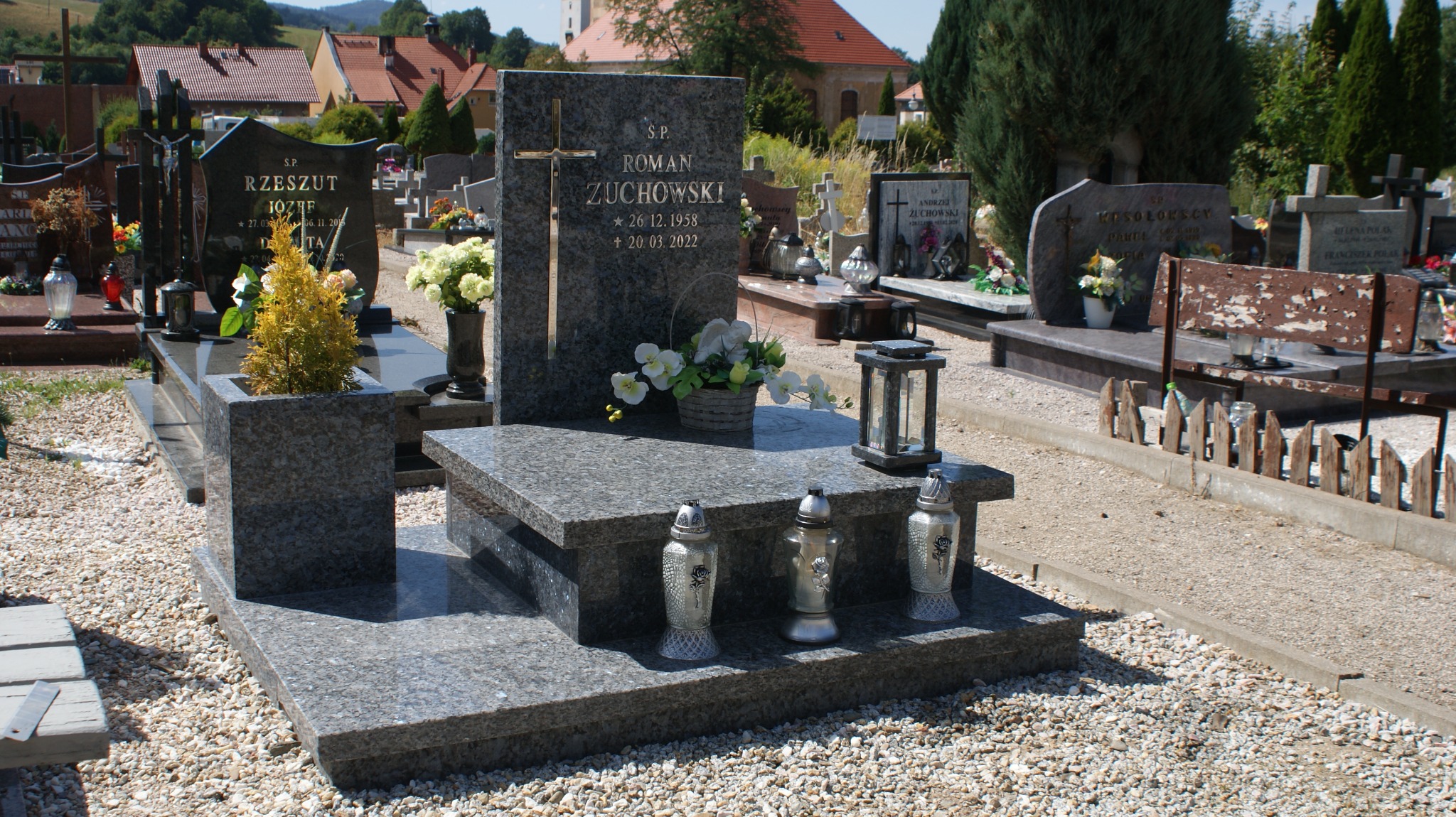 Granitowy pomnik z wygrawerowanym krzyżem i danymi zmarłego, ozdobiony kwiatami i lampionami na cmentarzu z innymi nagrobkami w tle.