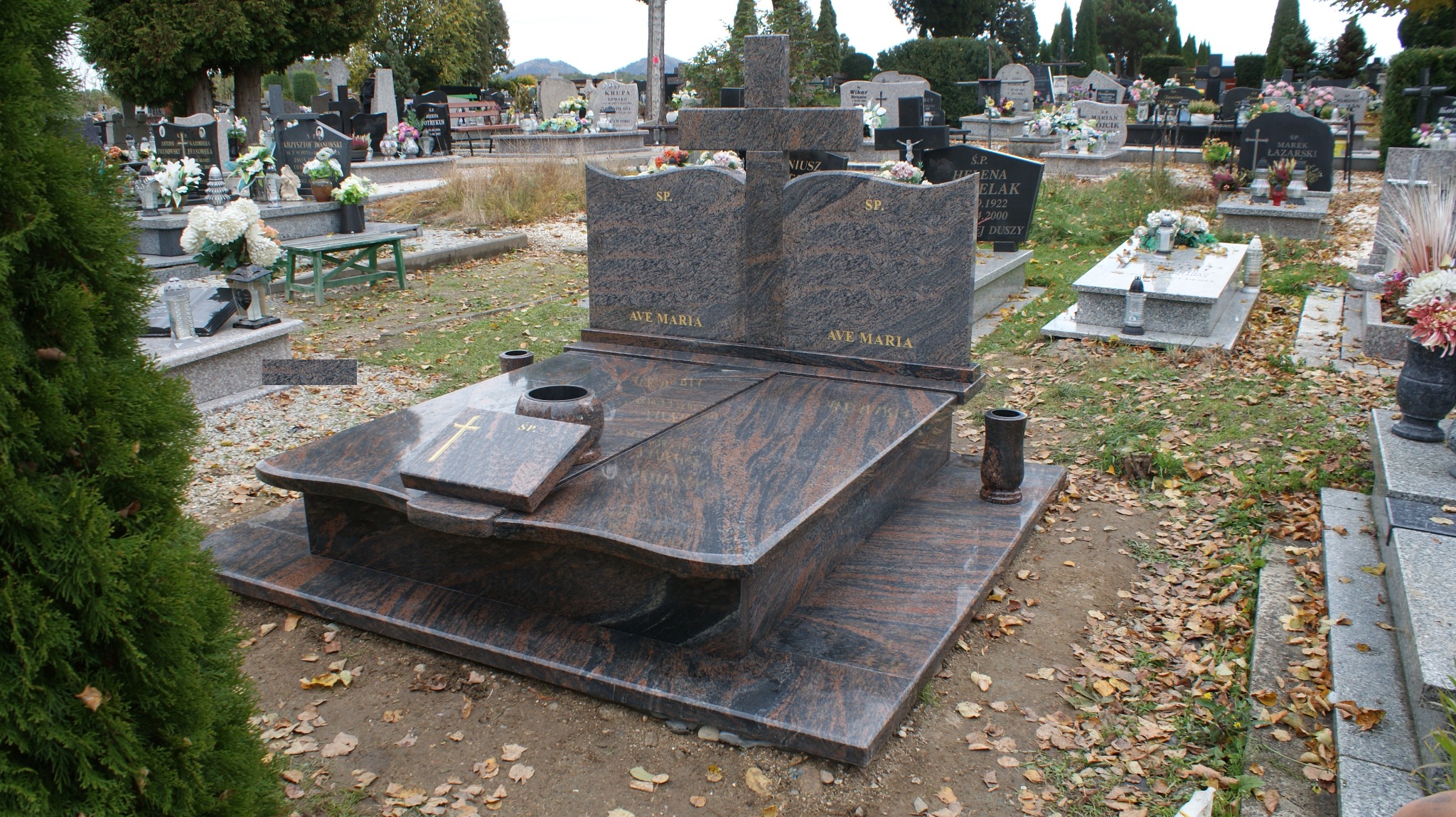 Nowoczesny, dwupoziomowy pomnik z ciemnego, wzorzystego kamienia z napisem 'AVE MARIA', umieszczony na cmentarzu z widocznymi innymi nagrobkami i opadłymi liśćmi.