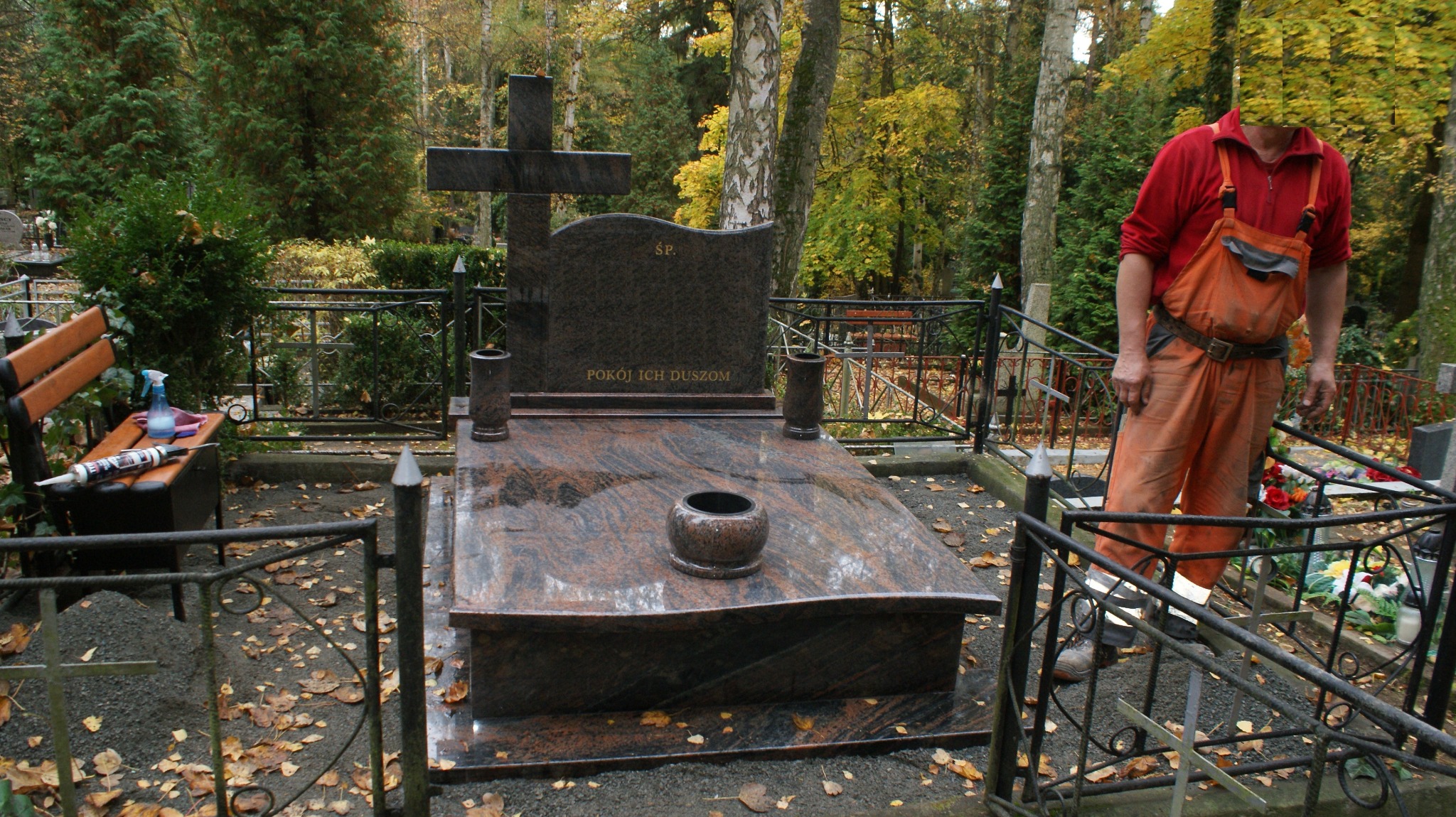 Polerowany, brązowo-czarny pomnik z krzyżem i napisem 'Ś.P. Pokój Ich Duszom', otoczony metalowym ogrodzeniem, z pracownikiem w pomarańczowych spodniach i czerwonej koszuli w tle, na cmentarzu...