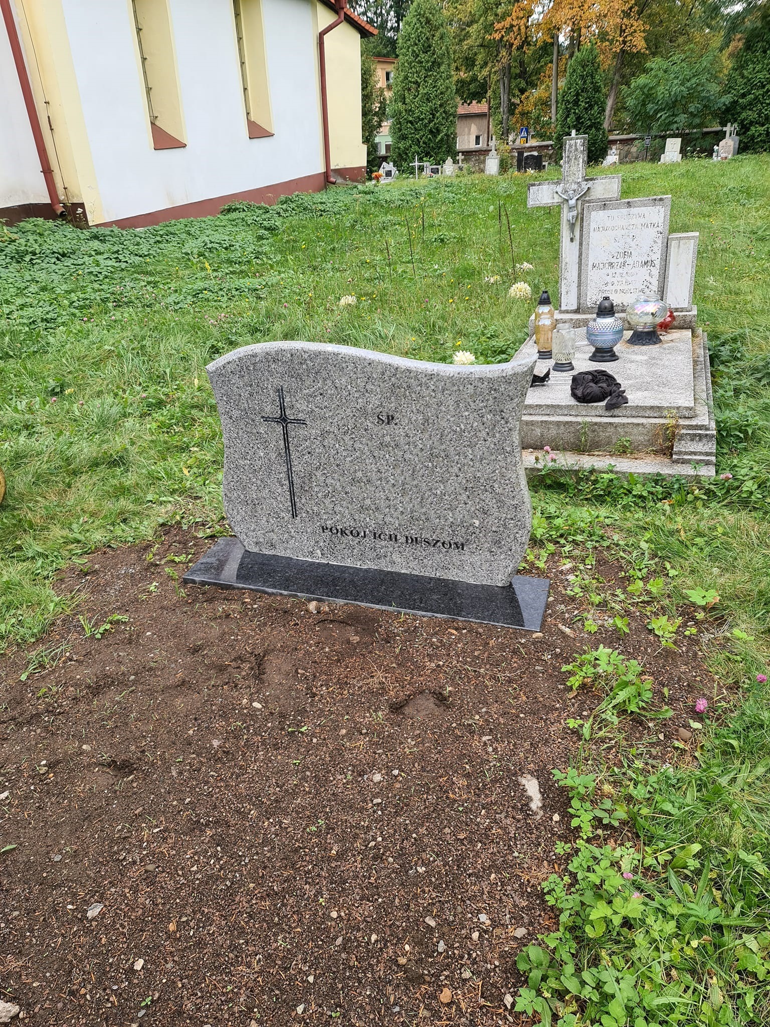 Nowy, szary nagrobek z krzyżem i napisem 'Pokój Ich Duszom' na czarnym cokole, świeżo posadowiony na ziemi w otoczeniu trawy i innych nagrobków na cmentarzu.