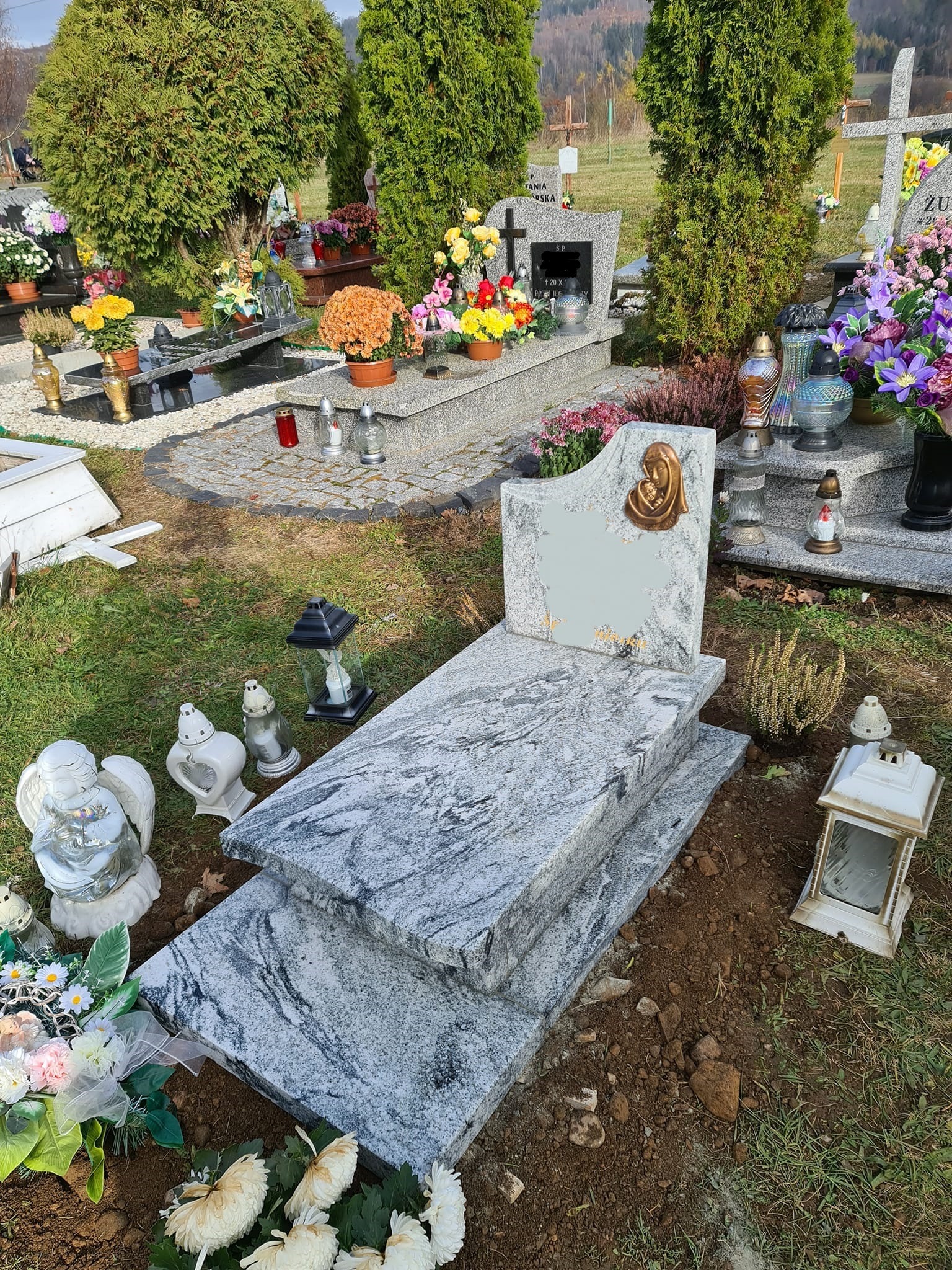Jasny, marmurkowy pomnik z płaskorzeźbą Matki Boskiej, otoczony lampionami i kwiatami na cmentarzu, z widoczną ziemią wokół podstawy.