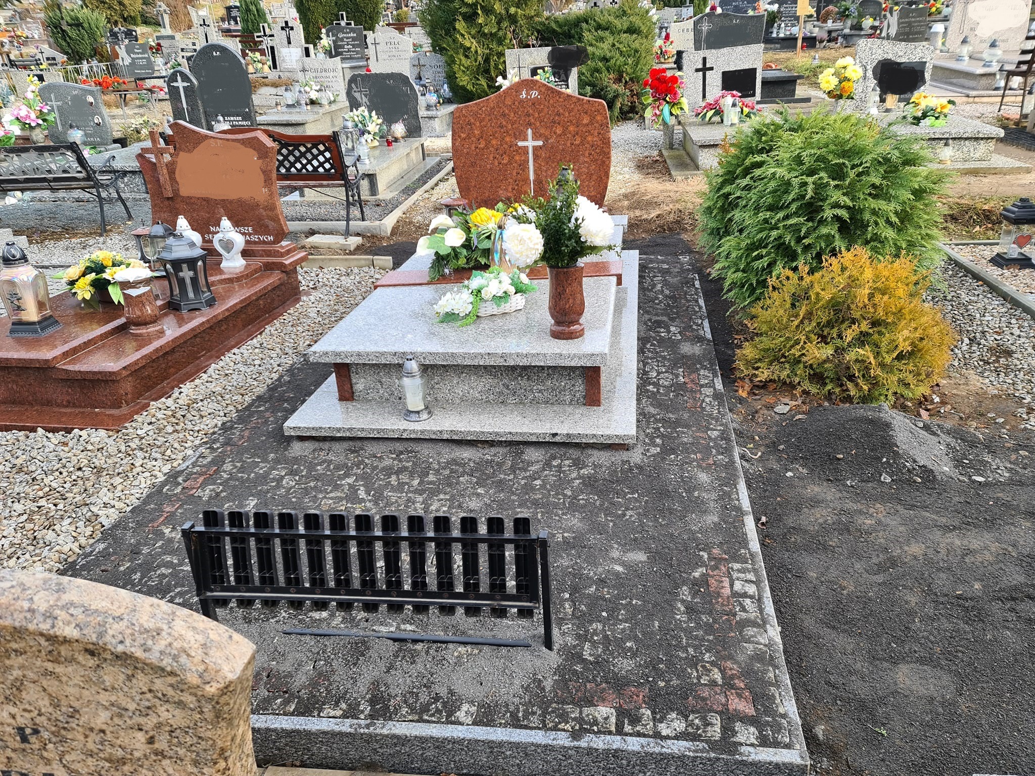 Granitowy nagrobek z brązowym akcentem i krzyżem, ozdobiony kwiatami w wazonie i koszyku, na cmentarzu z licznymi grobami w tle, z czarnym metalowym płotkiem u podstawy.