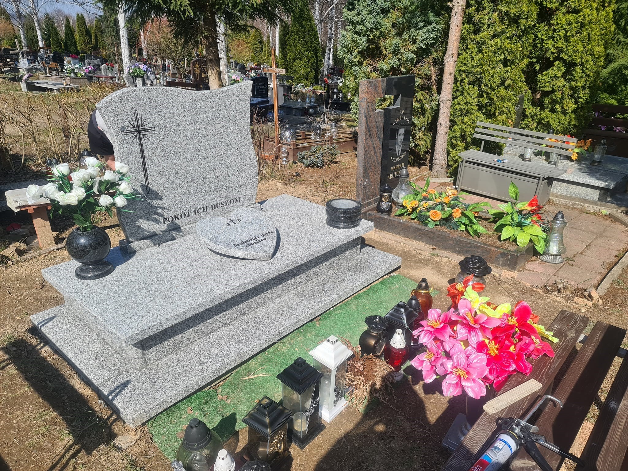 Jasnoszary, granitowy pomnik z napisem i krzyżem na cmentarzu, otoczony kwiatami w wazonach i lampionami, z drewnianą ławką i aplikatorem do silikonu w tle.