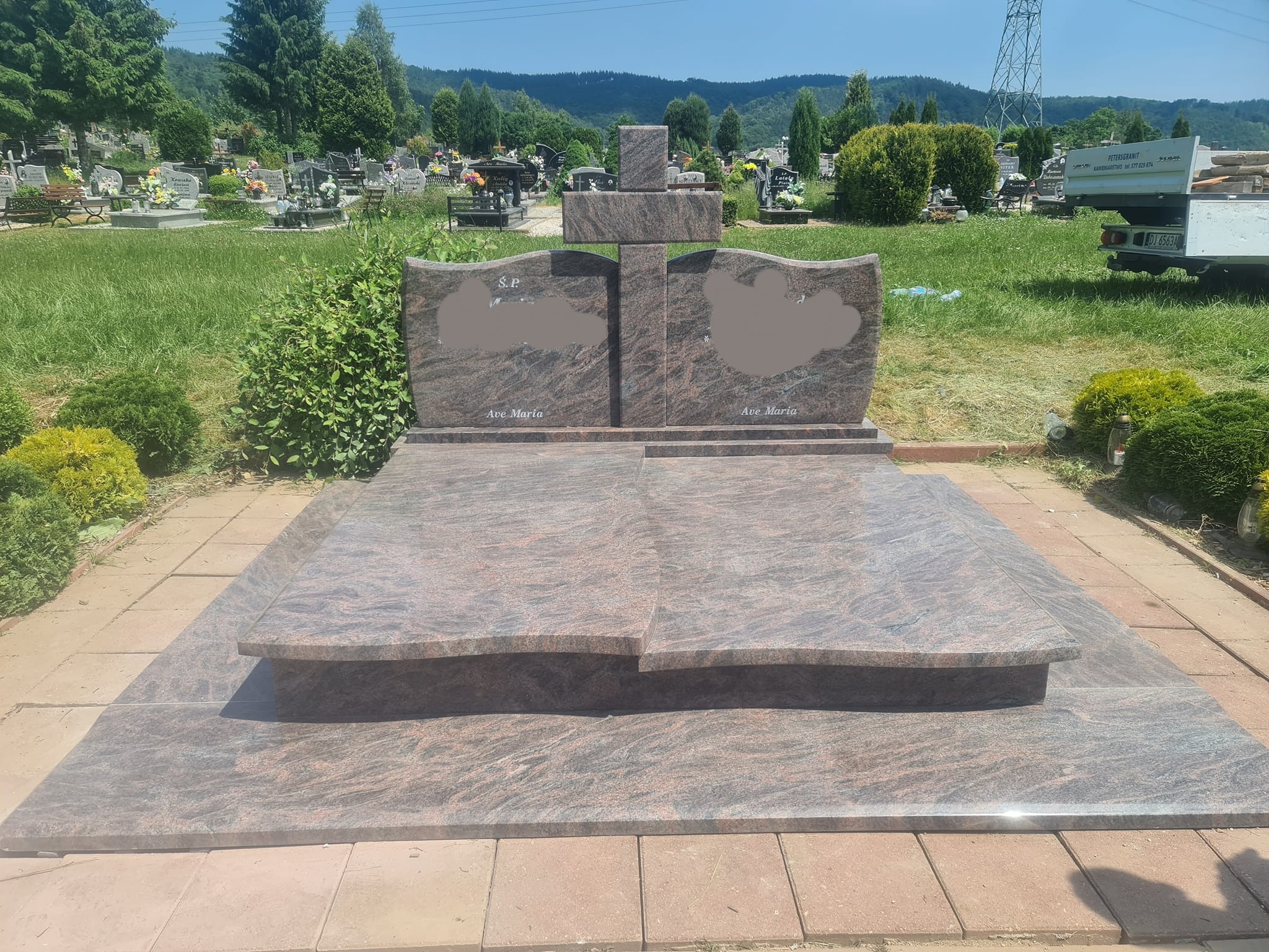 Granitowy pomnik z krzyżem na cmentarzu, z wygrawerowanymi napisami 'S.P.' i 'Ave Maria', otoczony zielenią i innymi nagrobkami w tle.