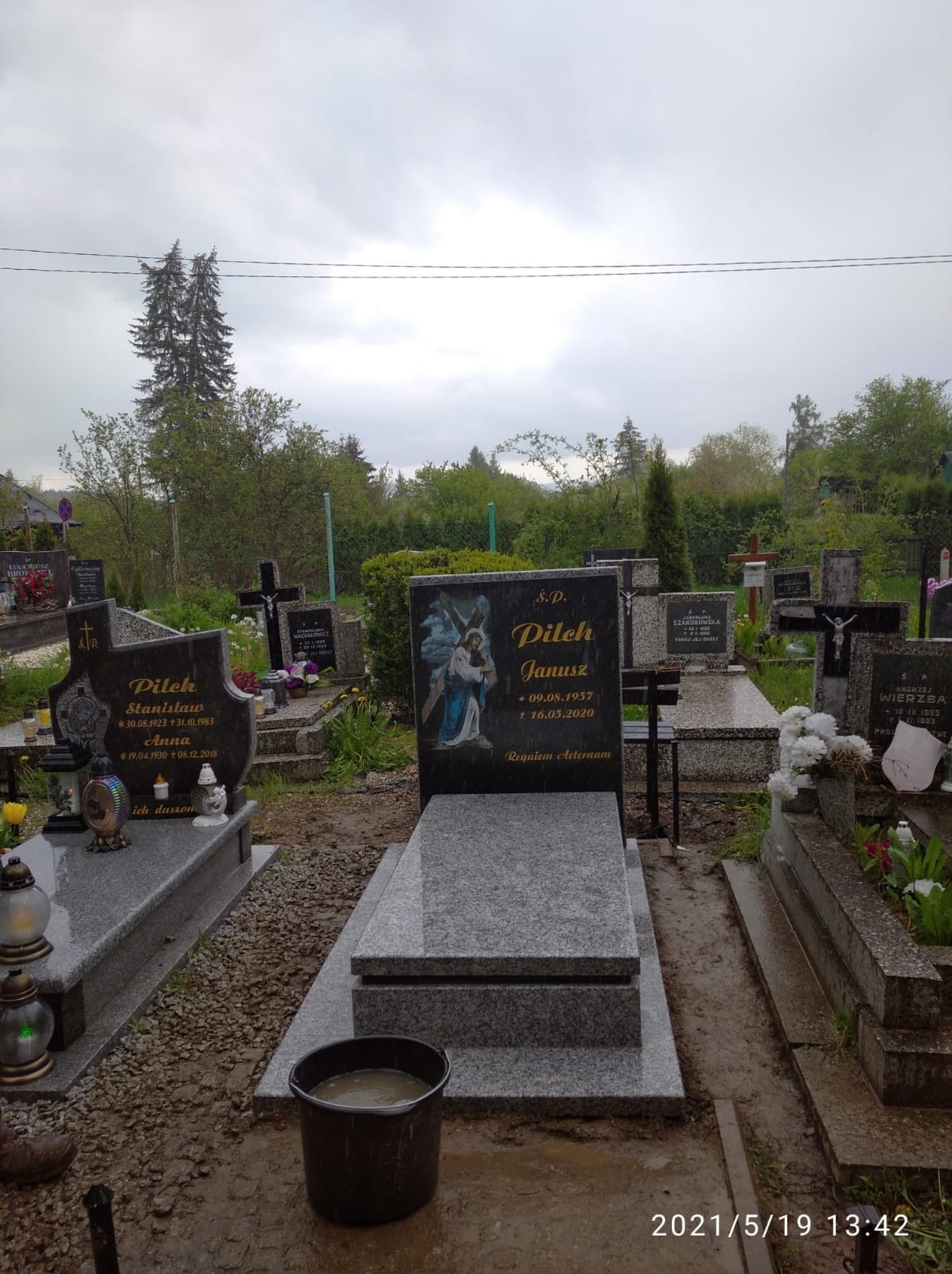 Nowy, szary, granitowy pomnik z czarną tablicą z wizerunkiem anioła i napisem, na cmentarzu w pochmurny dzień. Obok inne nagrobki i wiadro z wodą.