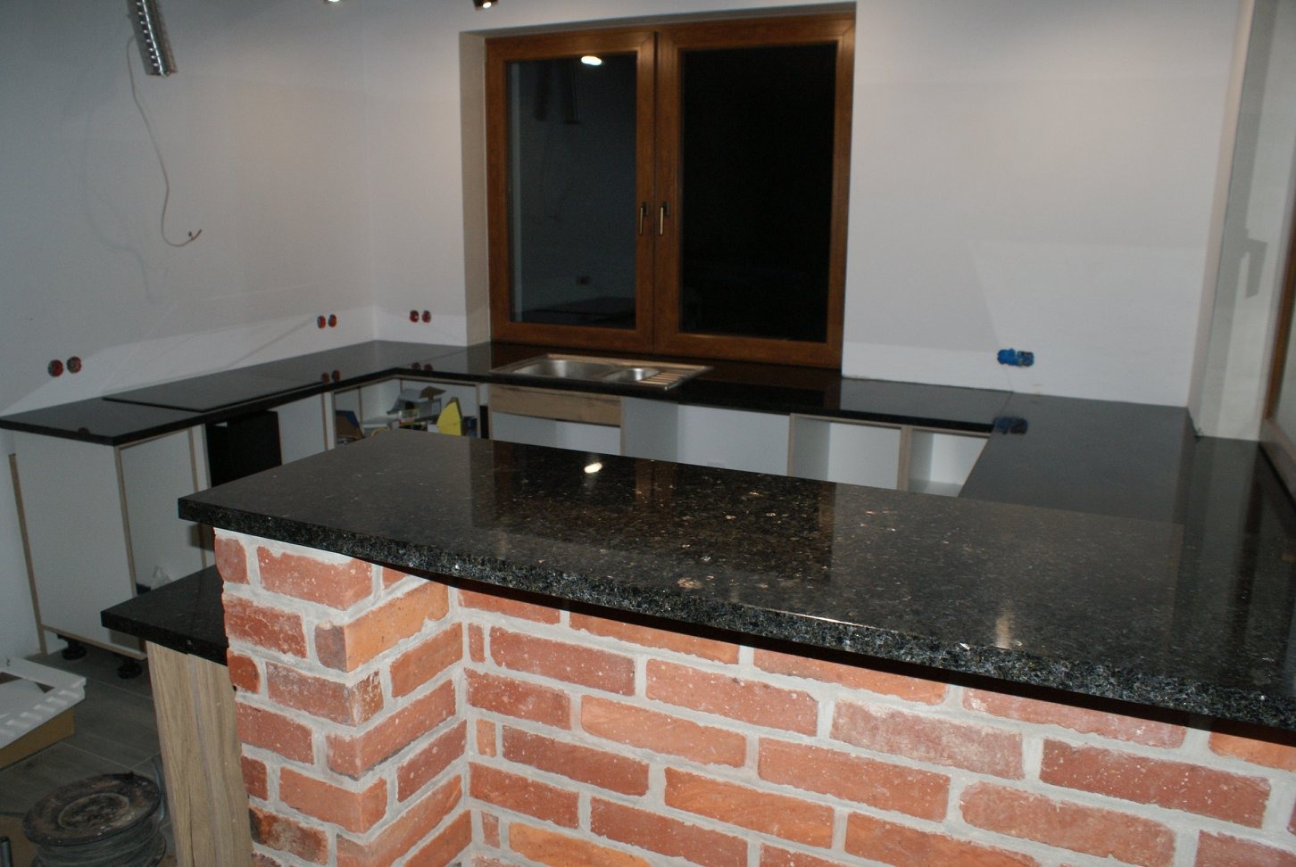 Czarny, błyszczący blat kamienny z widoczną strukturą, wsparty na murze z czerwonej cegły w kuchni w trakcie montażu; widoczne niedokończone szafki i instalacje.