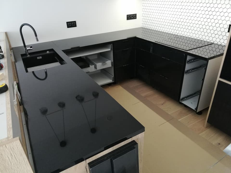Czarny, błyszczący blat kuchenny z zamontowanym zlewozmywakiem i baterią, widoczne szuflady w trakcie montażu, ściana wyłożona białą mozaiką heksagonalną.
