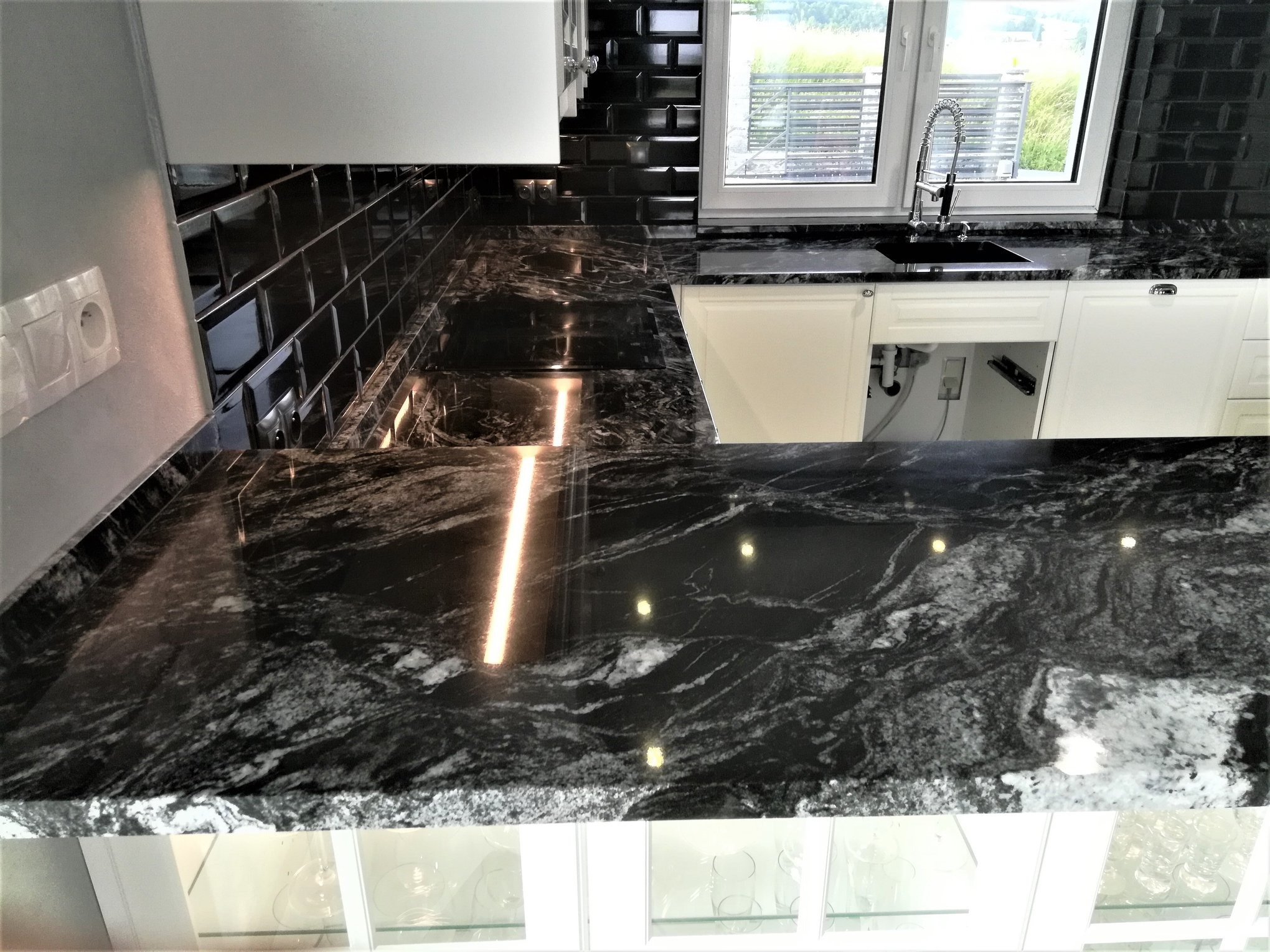 Czarny blat kuchenny z kamienia o wyraźnym, białym użyleniu, w nowoczesnej kuchni z czarnymi, błyszczącymi płytkami na ścianie i białymi szafkami, widok na zewnątrz przez okno.