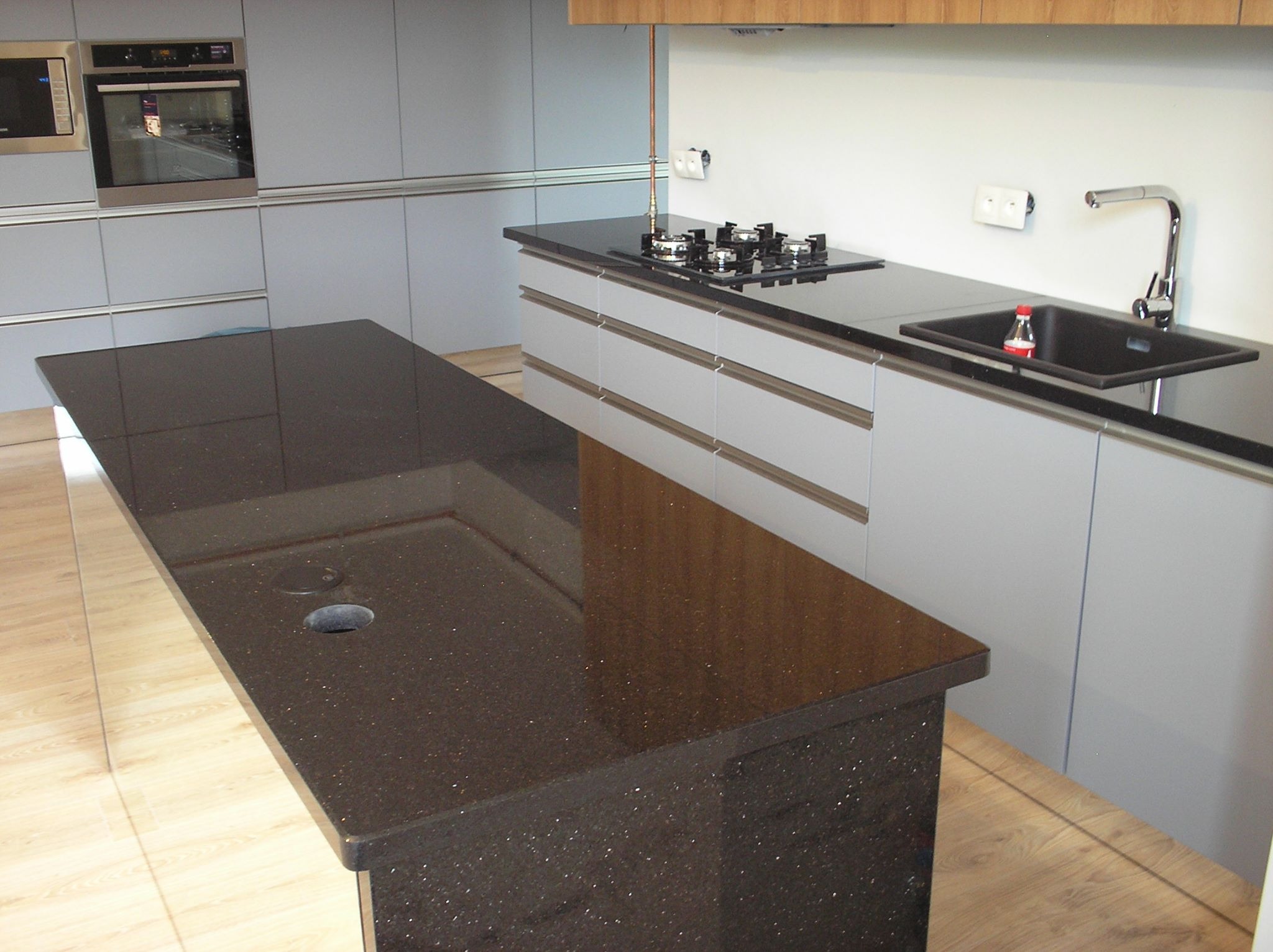 Błyszczący, ciemny blat z konglomeratu w nowoczesnej kuchni z szarymi szafkami, widoczna płyta grzewcza i zlew z baterią, butelka płynu w zlewie.