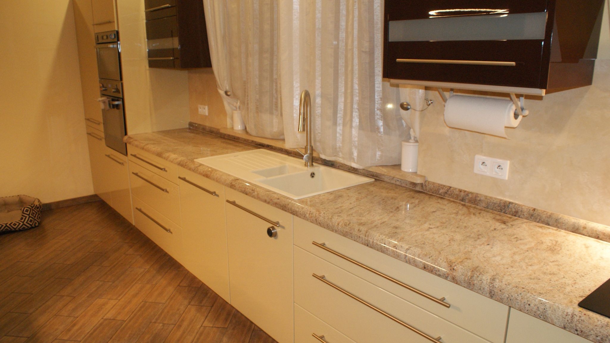 Beżowy blat kuchenny z delikatnym użyleniem, zintegrowany z białym zlewozmywakiem, nowoczesne szafki z podłużnymi uchwytami, drewniana podłoga i okno z jasną zasłoną w tle.