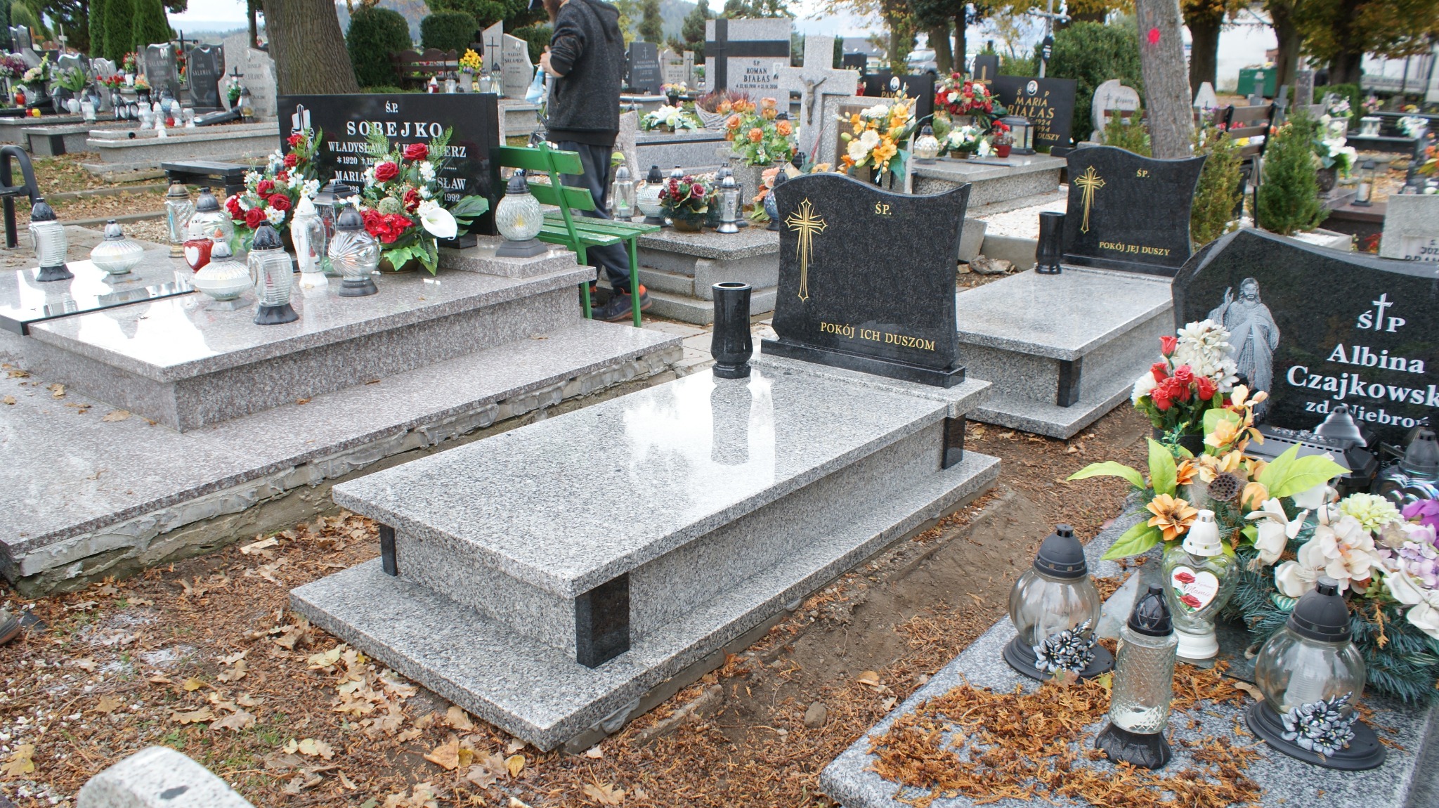 Szary granitowy nagrobek z czarną wazą i tablicą z napisem 'Pokój Ich Duszom' na cmentarzu, widok z perspektywy lekko z góry, jesienne liście na ziemi.