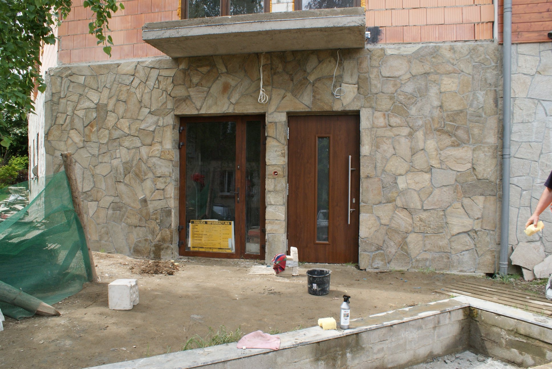 Wykończenie elewacji budynku kamieniem naturalnym w trakcie prac, widoczne narzędzia i materiały budowlane, okno z tablicą informacyjną, brązowe drzwi wejściowe.