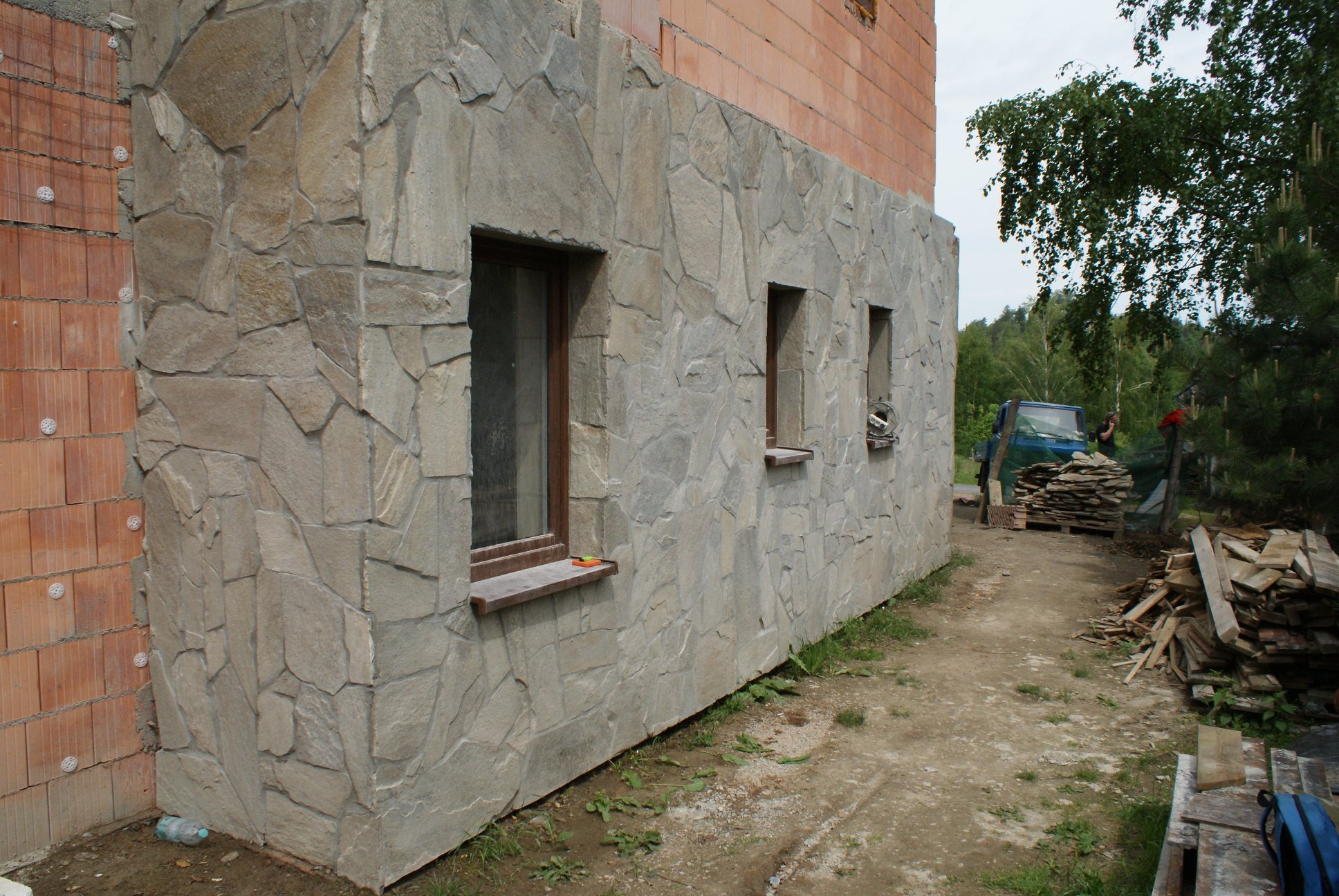 Elewacja budynku w trakcie wykańczania, pokryta nieregularnym kamieniem naturalnym w odcieniach szarości, z widocznymi trzema oknami z drewnianymi parapetami, ceglana ściana w tle, oraz stos...