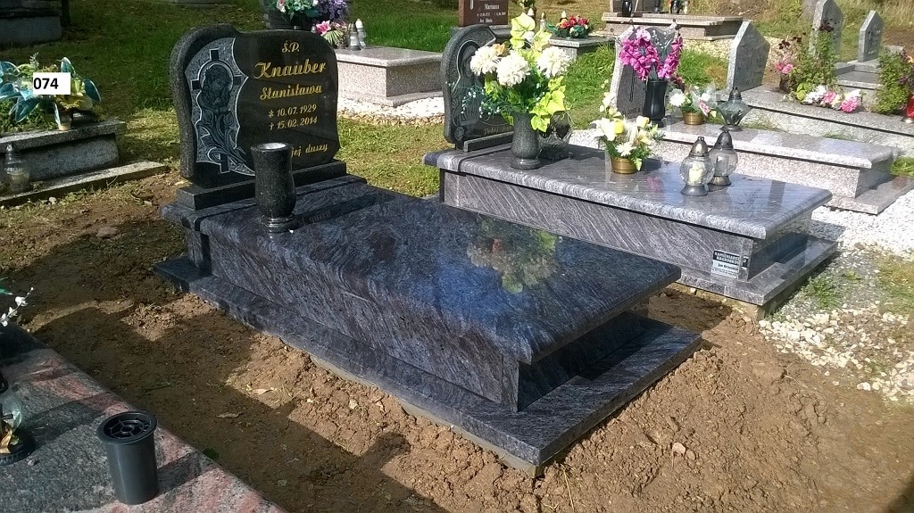 Granitowy pomnik z ciemnym blatem i wygrawerowanym wizerunkiem postaci, otoczony ziemią, z kwiatami w wazonie i zniczami na sąsiednim grobie.