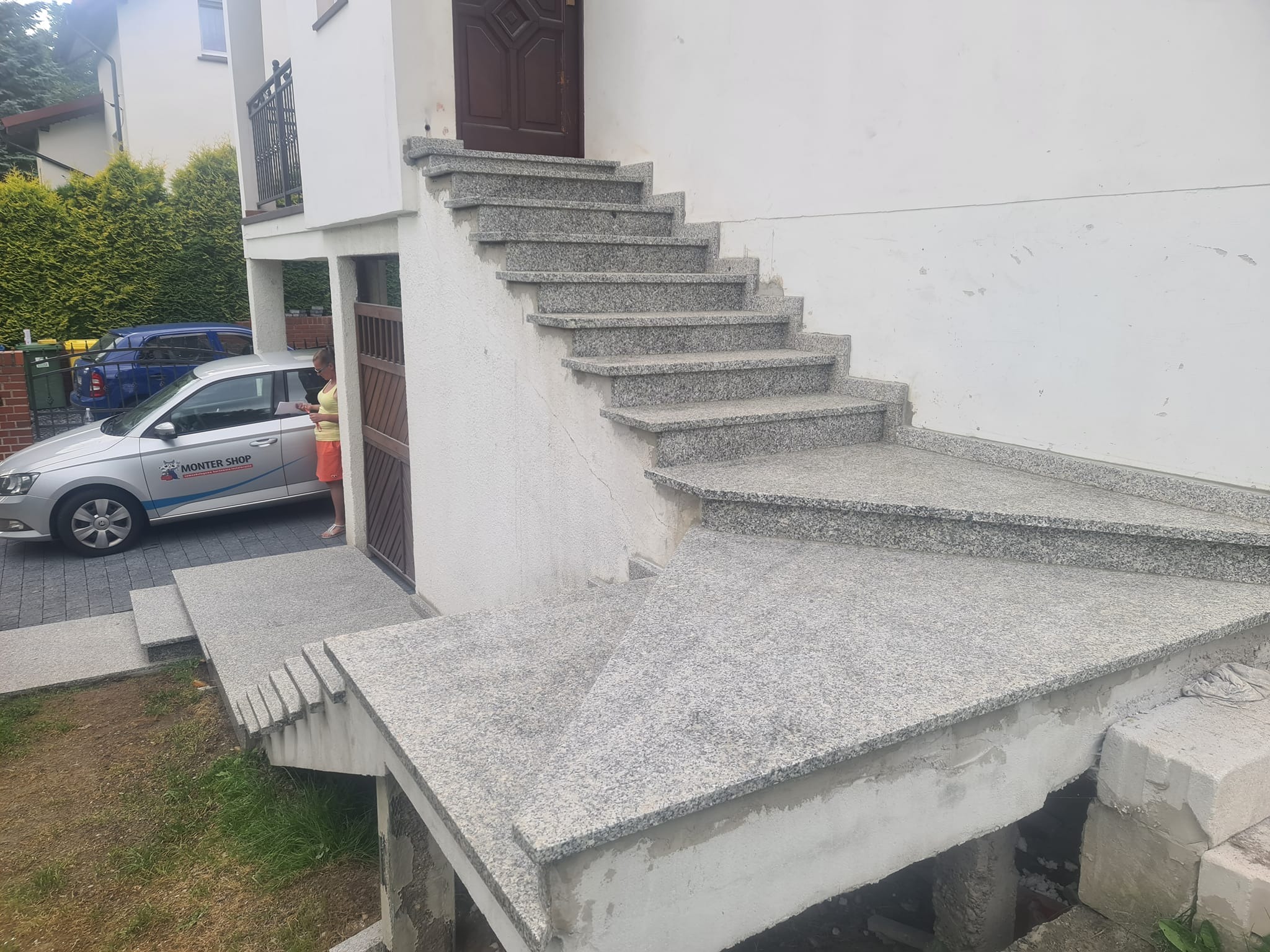 Zewnętrzne schody wejściowe do domu, wykończone jasnym, nakrapianym kamieniem, widoczne niedokończone prace pod schodami, samochód z logo firmy w tle po lewej stronie.