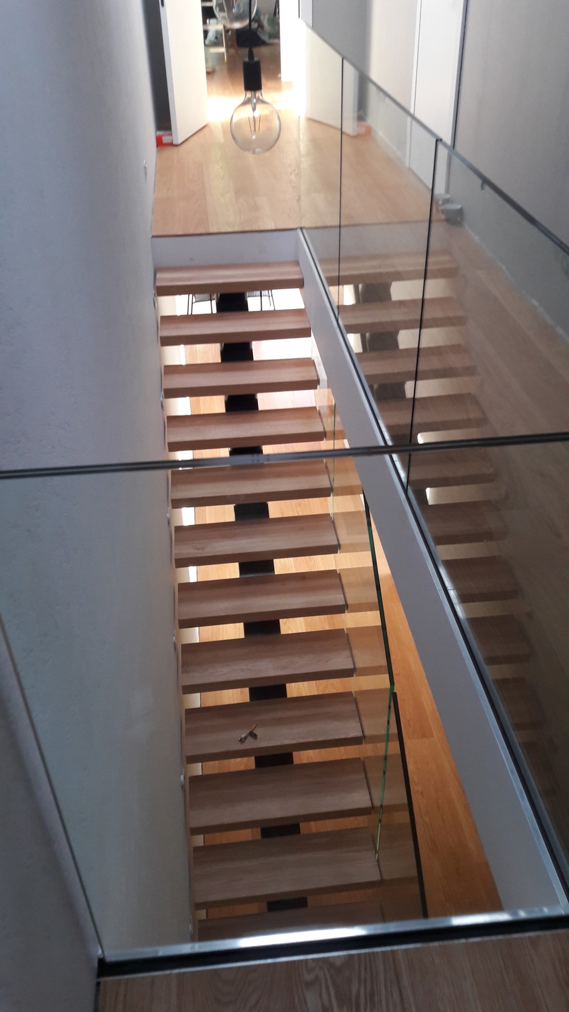 Nowoczesne schody z drewnianymi stopniami i szklaną balustradą, widok z góry, z minimalistyczną lampą w tle.