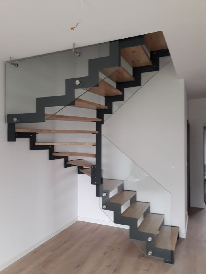 Nowoczesne schody z drewnianymi stopniami i szarą metalową konstrukcją, zabezpieczone szklaną balustradą, widoczne mocowania do ściany, minimalistyczny design.