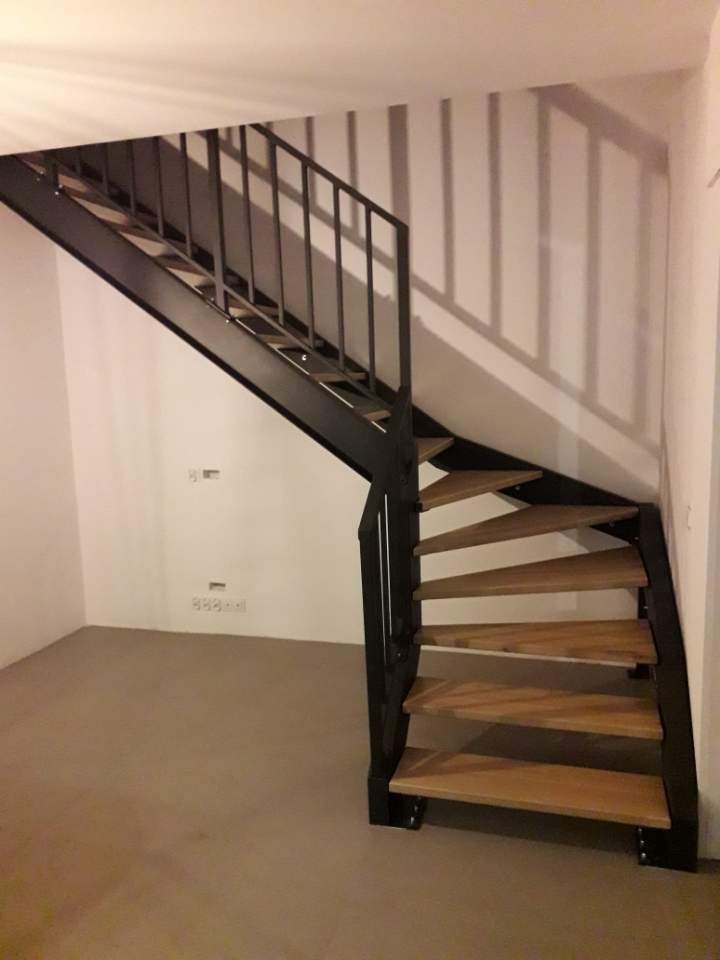 Metalowe schody spiralne z drewnianymi stopniami i czarną balustradą w surowym wnętrzu, rzucające geometryczny cień na ścianę.