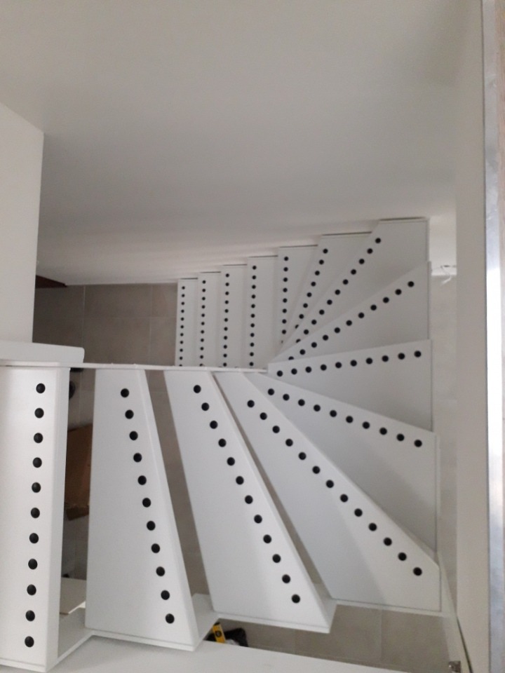 Białe schody spiralne z metalową konstrukcją i ozdobnymi czarnymi otworami, widok z dołu.