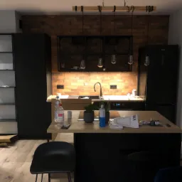 Nowoczesna kuchnia z ceglaną ścianą, czarnymi szafkami i wyspą z drewnianym blatem, na której stoją butelki z wodą i rośliny w doniczkach. Nad blatem wiszą lampy w stylu industrialnym.