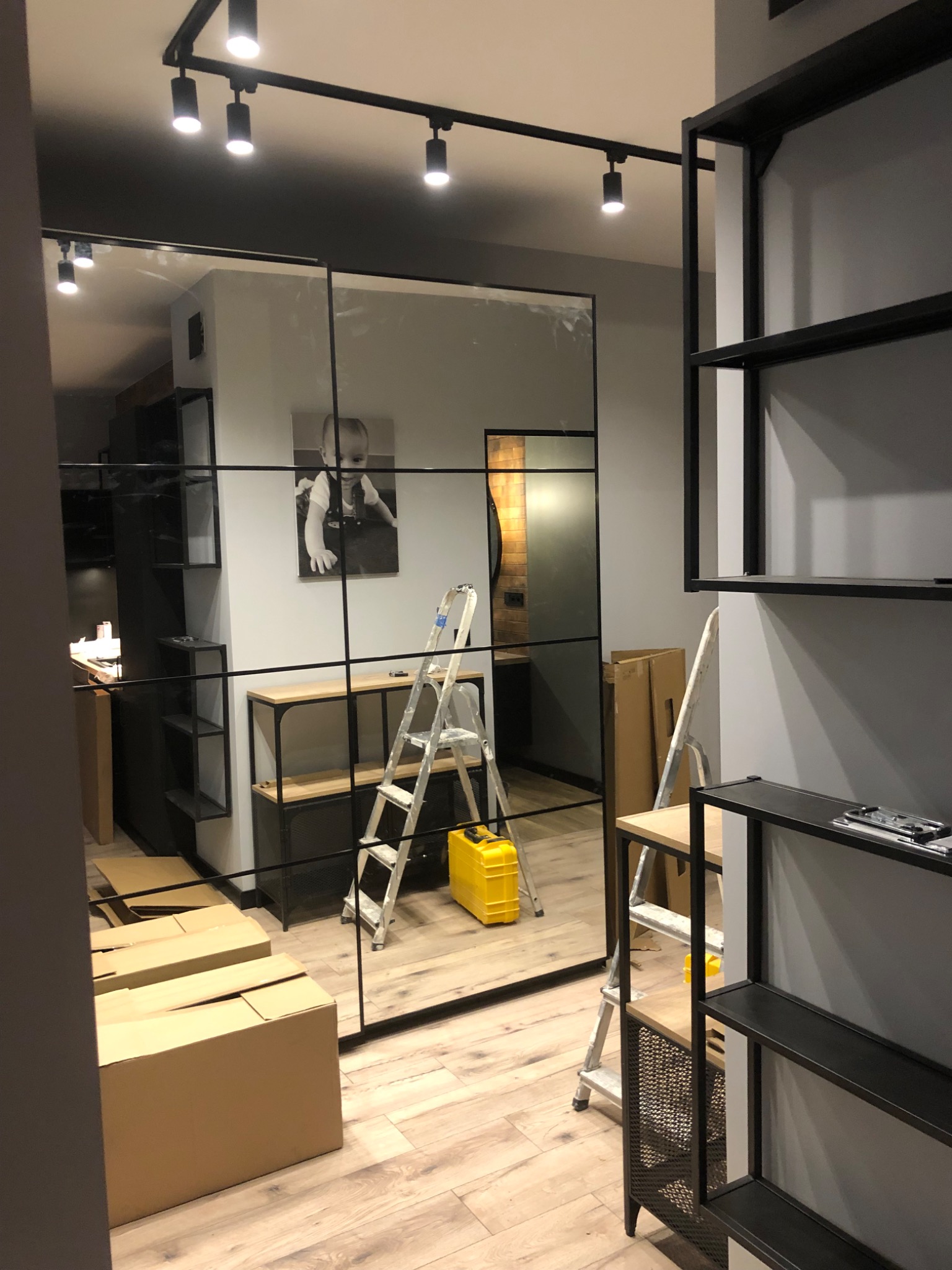 Montaż szklanej ściany działowej z czarnymi profilami w nowoczesnym wnętrzu, widoczne kartony, drabina i oświetlenie punktowe.