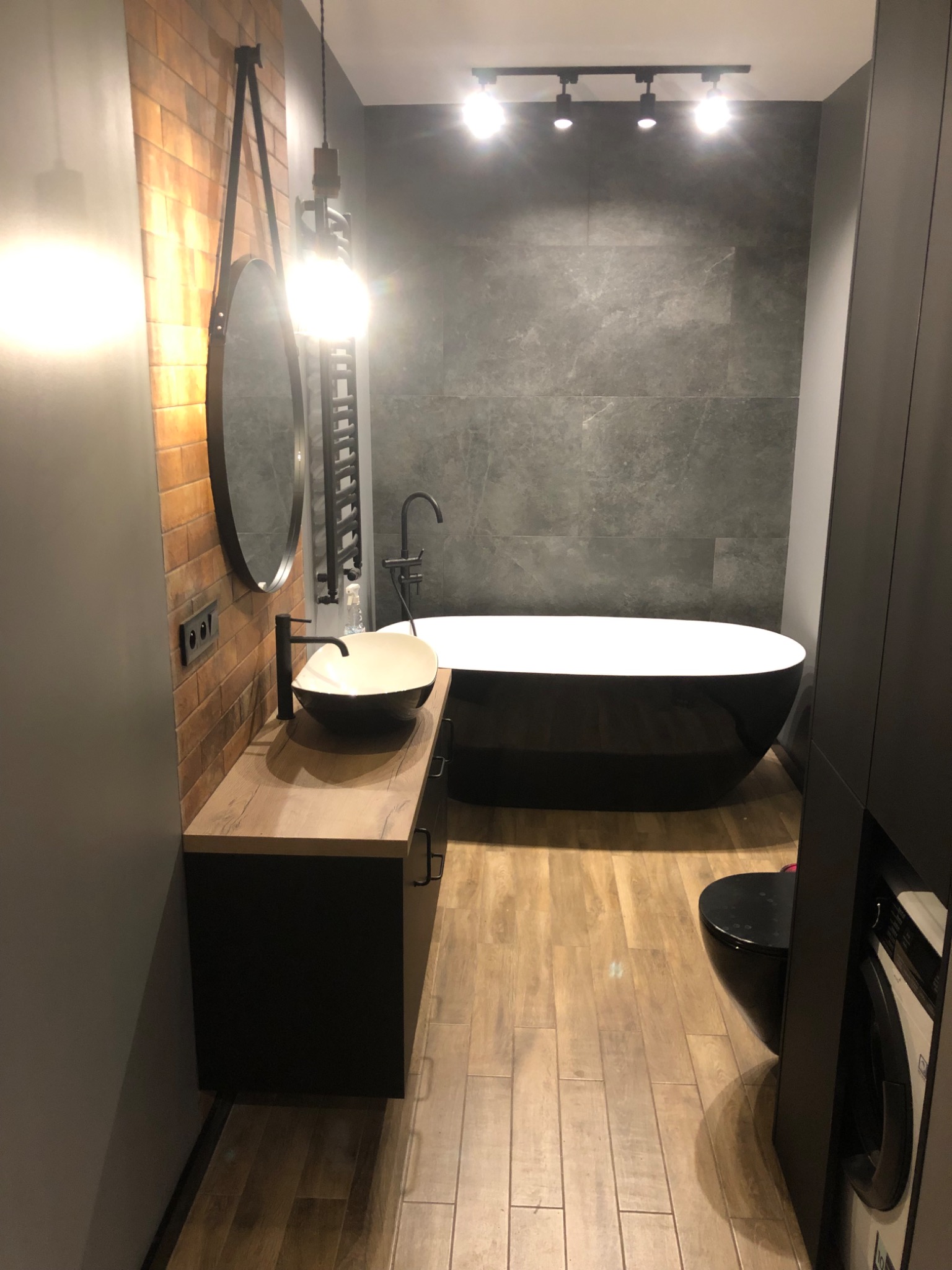 Nowoczesna łazienka z wanną wolnostojącą, umywalką nablatową w kształcie misy, podłogą imitującą drewno i ścianą z cegiełek.