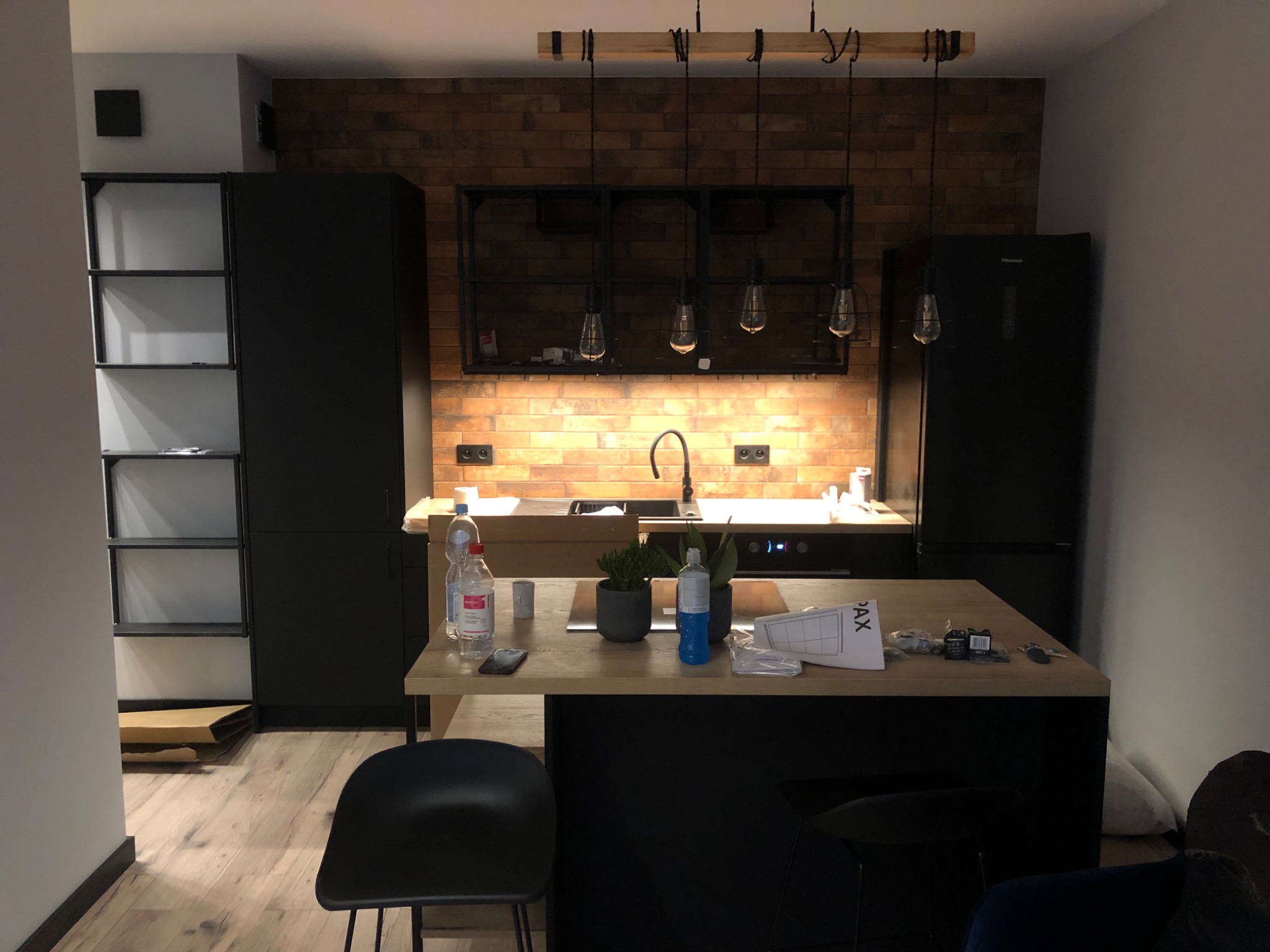 Nowoczesna kuchnia z ceglaną ścianą, czarnymi szafkami i wyspą z drewnianym blatem, na której stoją butelki z wodą i rośliny w doniczkach. Nad blatem wiszą lampy w stylu industrialnym.