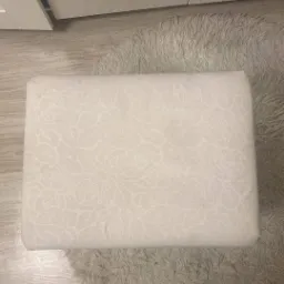 Pufa tapicerowana jasnym materiałem w subtelny wzór kwiatowy, leżąca na puszystym dywanie, widoczne stopy w skarpetkach z motywem misia, fragment szafy w tle.