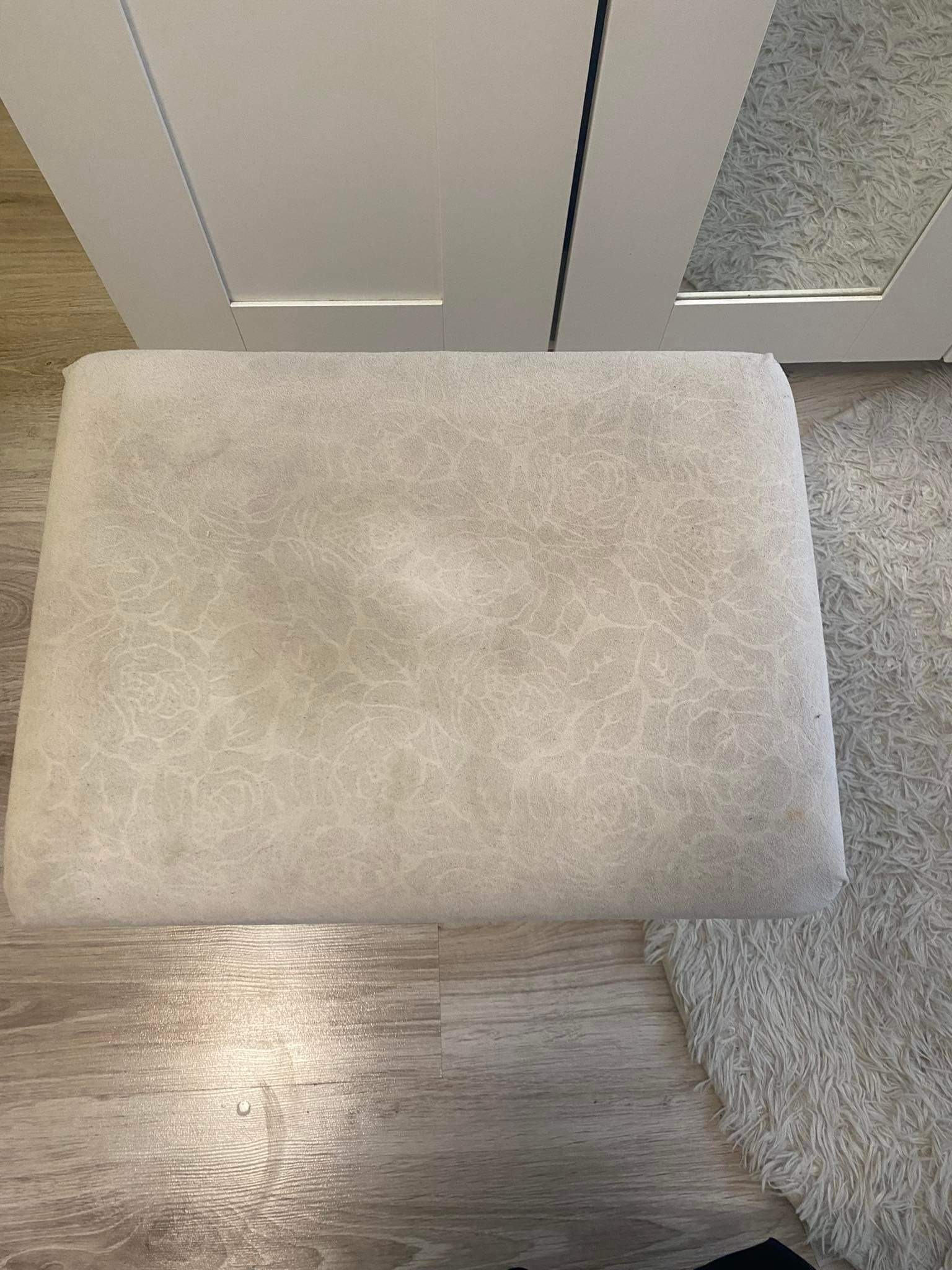 Tapicerowany puf w odcieniach beżu z delikatnym wzorem kwiatowym, ustawiony na panelach podłogowych obok puszystego dywanu, widoczne ślady użytkowania.