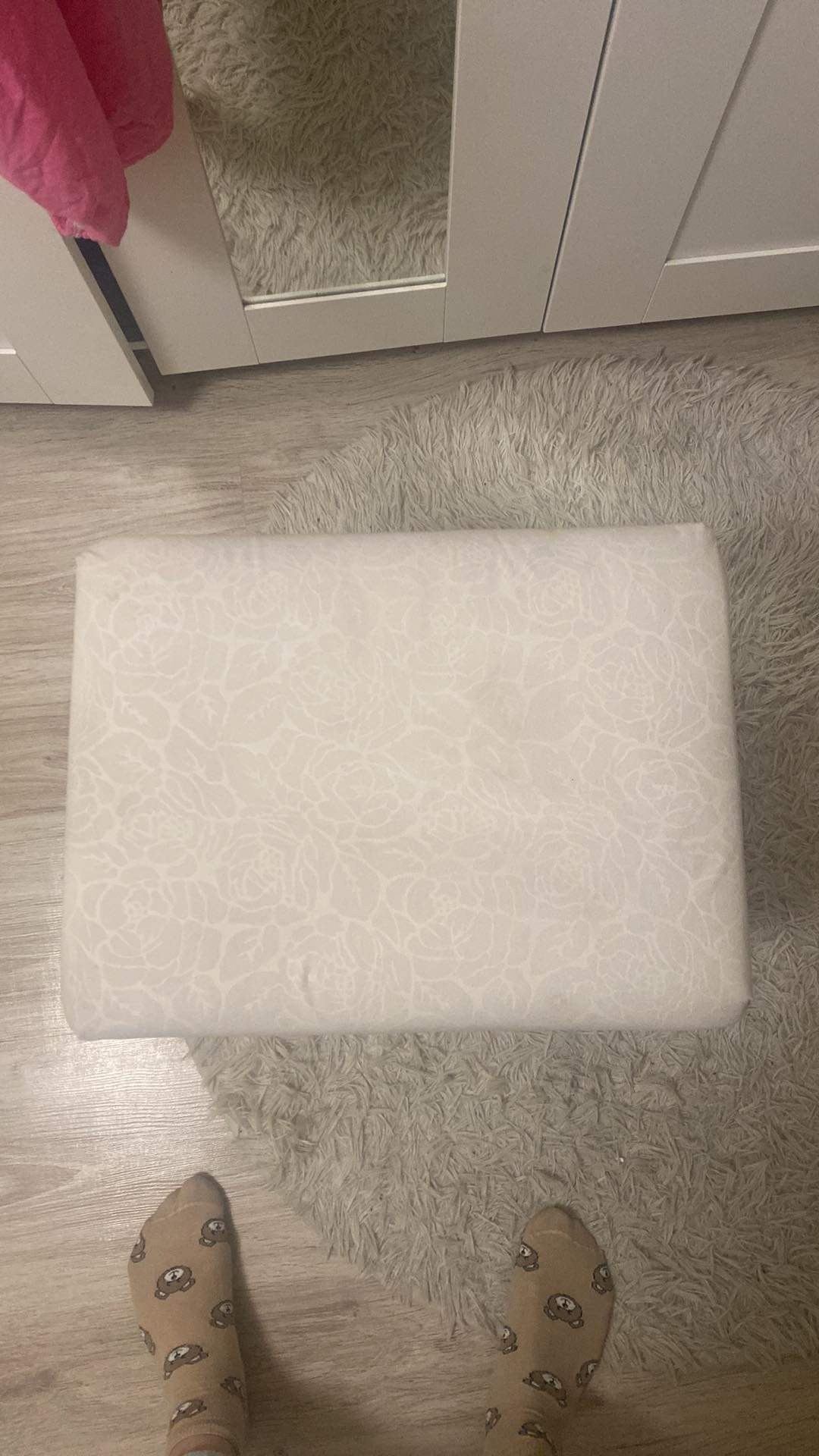 Pufa tapicerowana jasnym materiałem w subtelny wzór kwiatowy, leżąca na puszystym dywanie, widoczne stopy w skarpetkach z motywem misia, fragment szafy w tle.