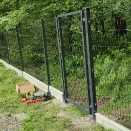 Nowoczesne, panelowe ogrodzenie z bramą w kolorze grafitowym, osadzone na betonowej podmurówce. W trawie leżą narzędzia i kartonowe pudełko.