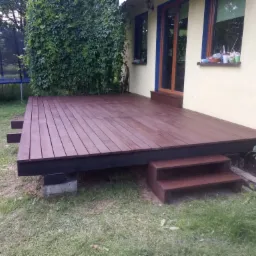 Nowo wybudowany drewniany taras przylegający do domu z żółtą elewacją, widoczne dwustopniowe schody wejściowe, pod tarasem betonowe podpory, w tle trawnik i trampolina.