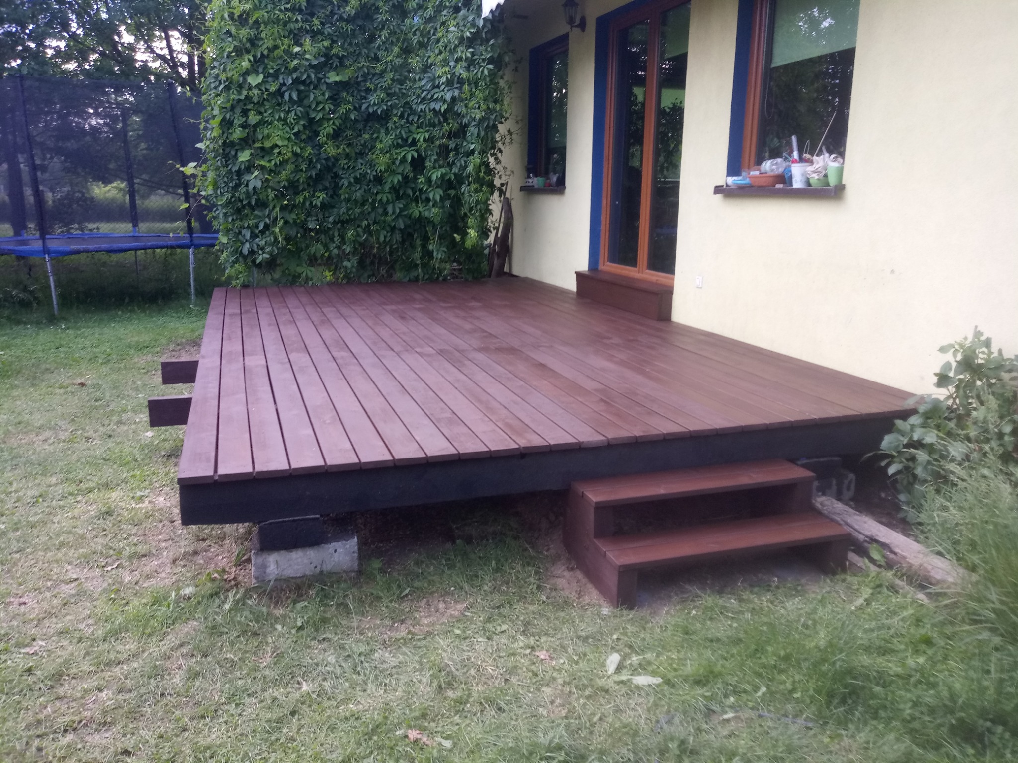 Nowo wybudowany drewniany taras przylegający do domu z żółtą elewacją, widoczne dwustopniowe schody wejściowe, pod tarasem betonowe podpory, w tle trawnik i trampolina.