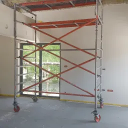 Wnętrze w trakcie prac wykończeniowych: rusztowanie na tle okna, ocieplone poddasze z widoczną konstrukcją metalową i drewnianą, surowa ściana z taśmą malarską przy podłodze.