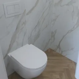 Podwieszana toaleta z zabudową na stelażu, wykończona płytkami imitującymi marmur, z drewnopodobną podłogą ułożoną w jodełkę, widoczne elementy instalacji przed zabudową.