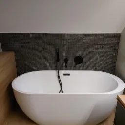 Nowoczesna łazienka z wolnostojącą białą wanną, czarna armatura i ściana wyłożona ciemnymi, pionowymi płytkami imitującymi drewno, oświetlenie górne.