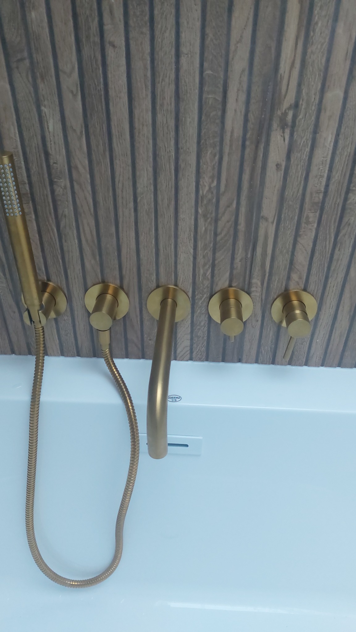 Złota armatura łazienkowa: bateria wannowa z deszczownicą i pokrętłami na tle ściany z drewnopodobnych paneli.