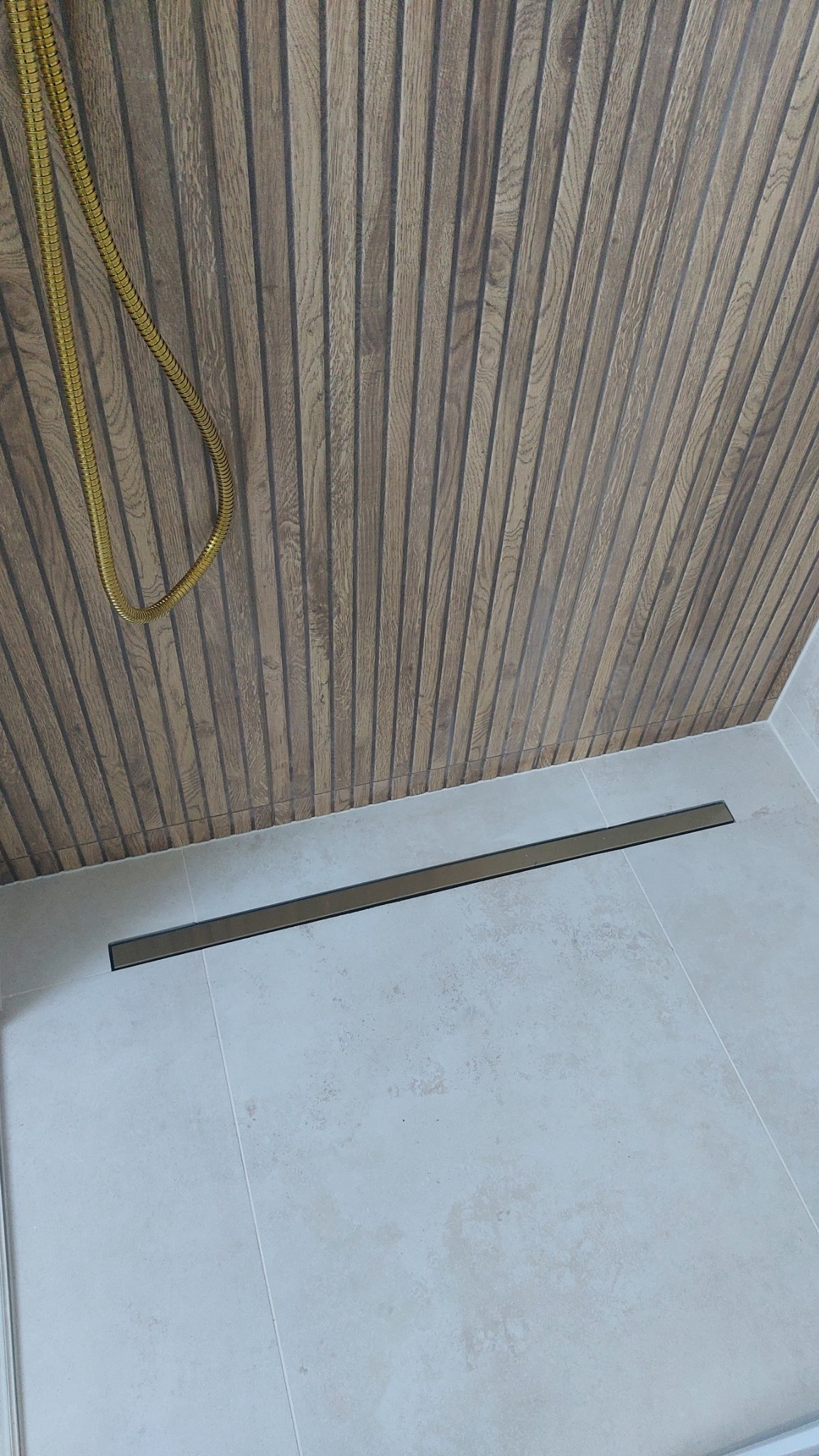 Nowoczesna kabina prysznicowa z drewnopodobną ścianą, złotym wężem prysznicowym i liniowym odpływem w podłodze z jasnych płytek.