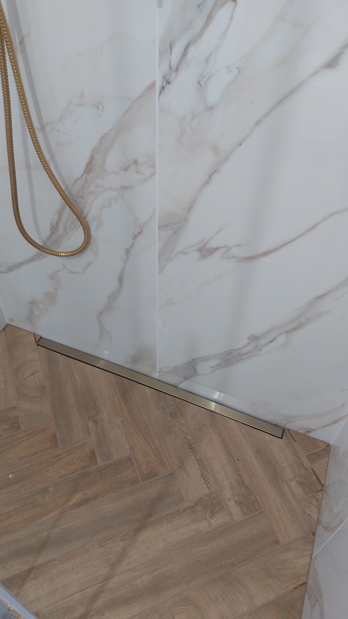 Detal łazienki z marmurowymi płytkami ściennymi i drewnopodobną podłogą ułożoną w jodełkę; widoczny odpływ liniowy w kolorze złotym oraz złoty wąż prysznicowy.